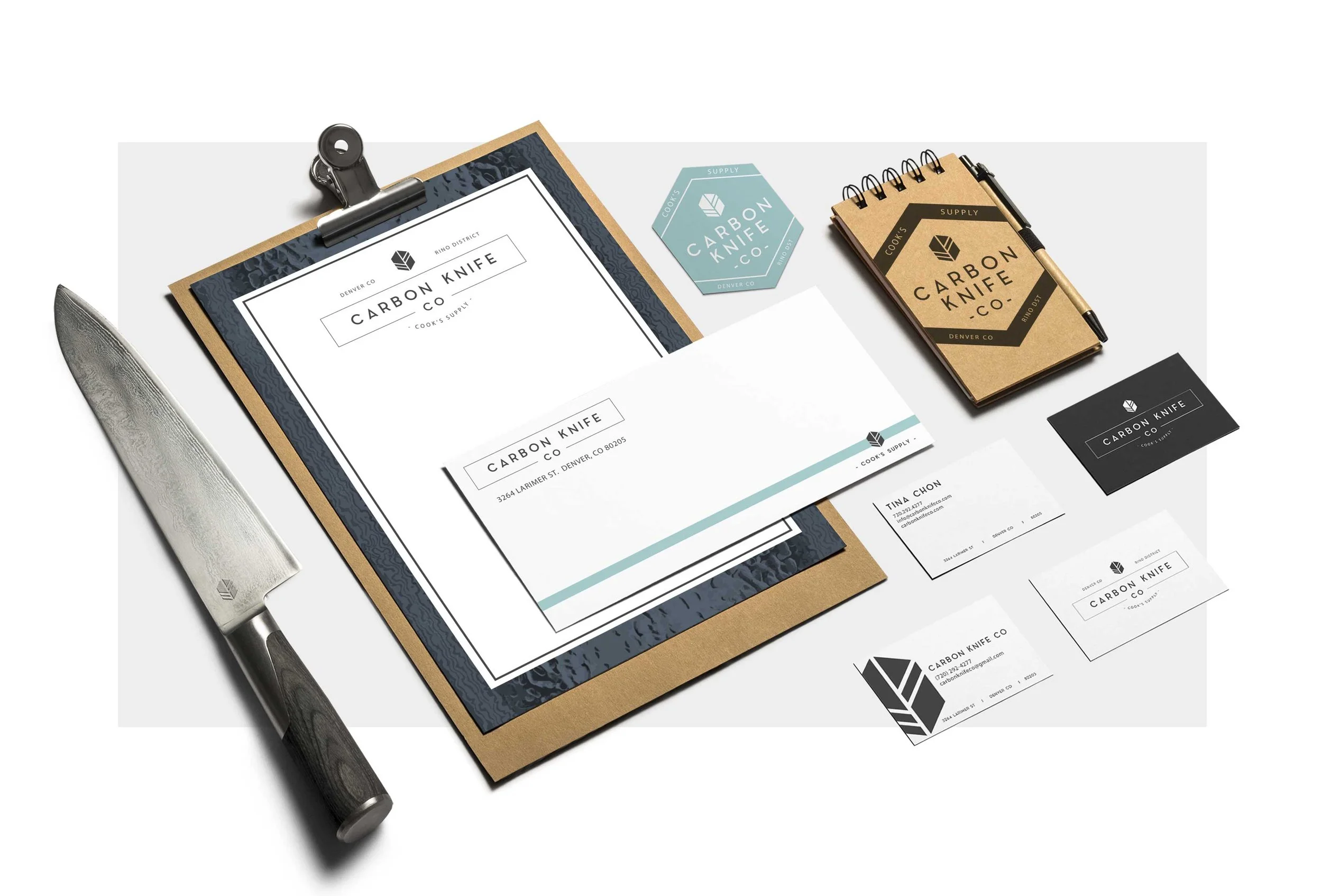 A workspace with a knife, business cards, a clipboard with a sheet of paper, a small notebook, and business stickers, all branded with 'Carbon Knife Co.' logo.