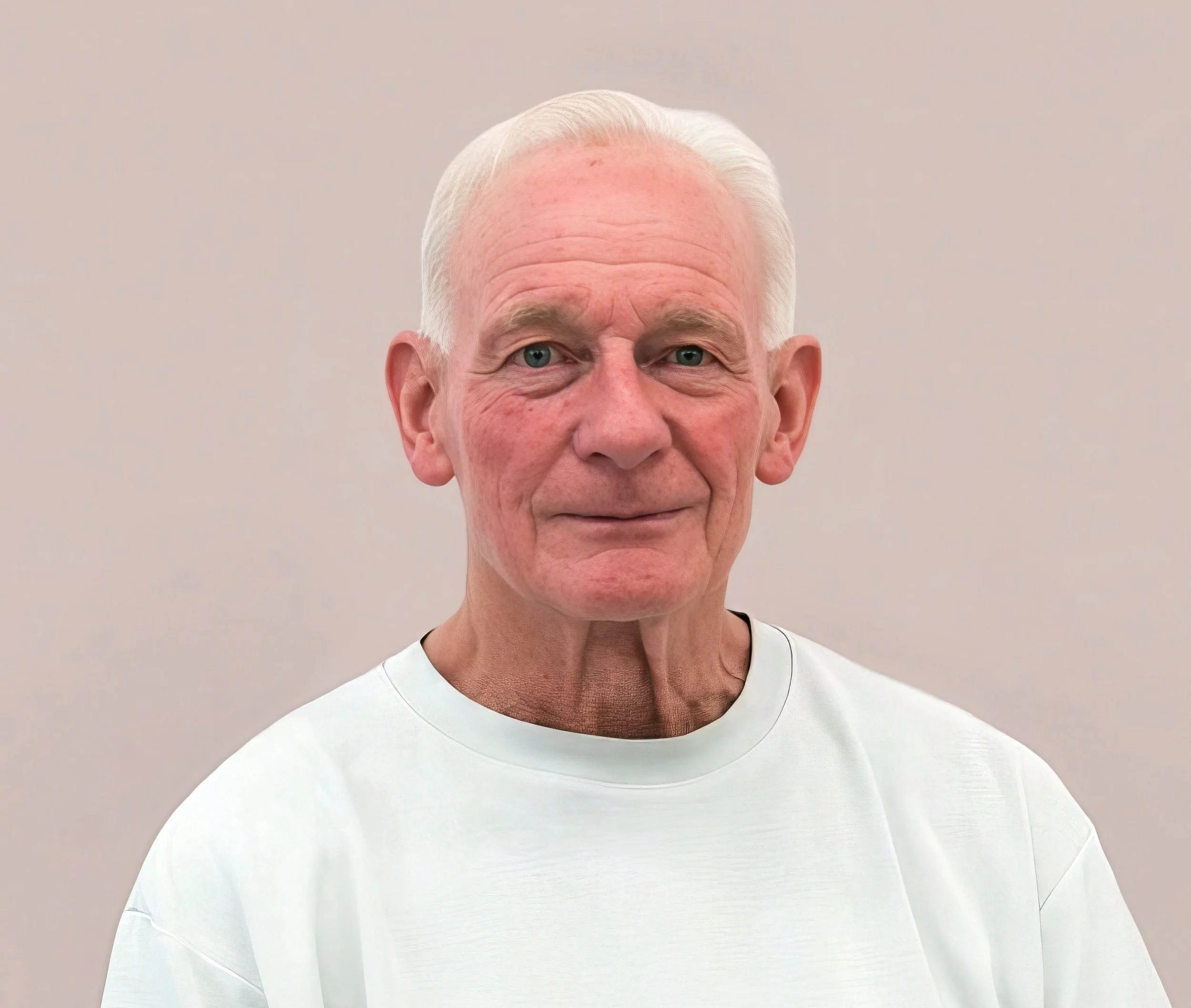 Close-up of a man with white hair, wearing a white shirt, standing against a plain light-colored background.