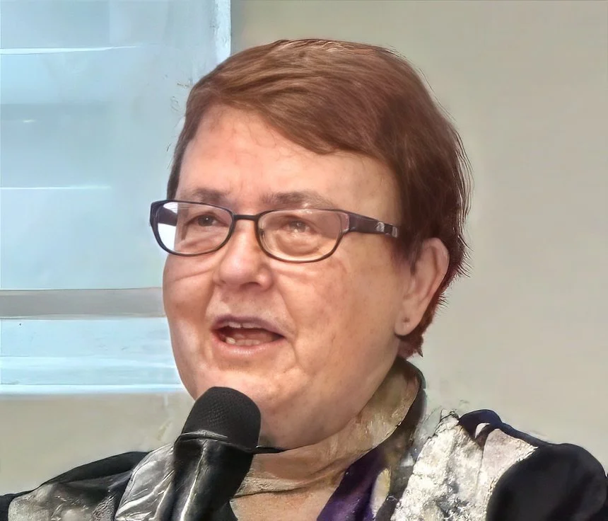 A woman with brown hair, glasses, and light skin, speaking into a microphone indoors near a window.