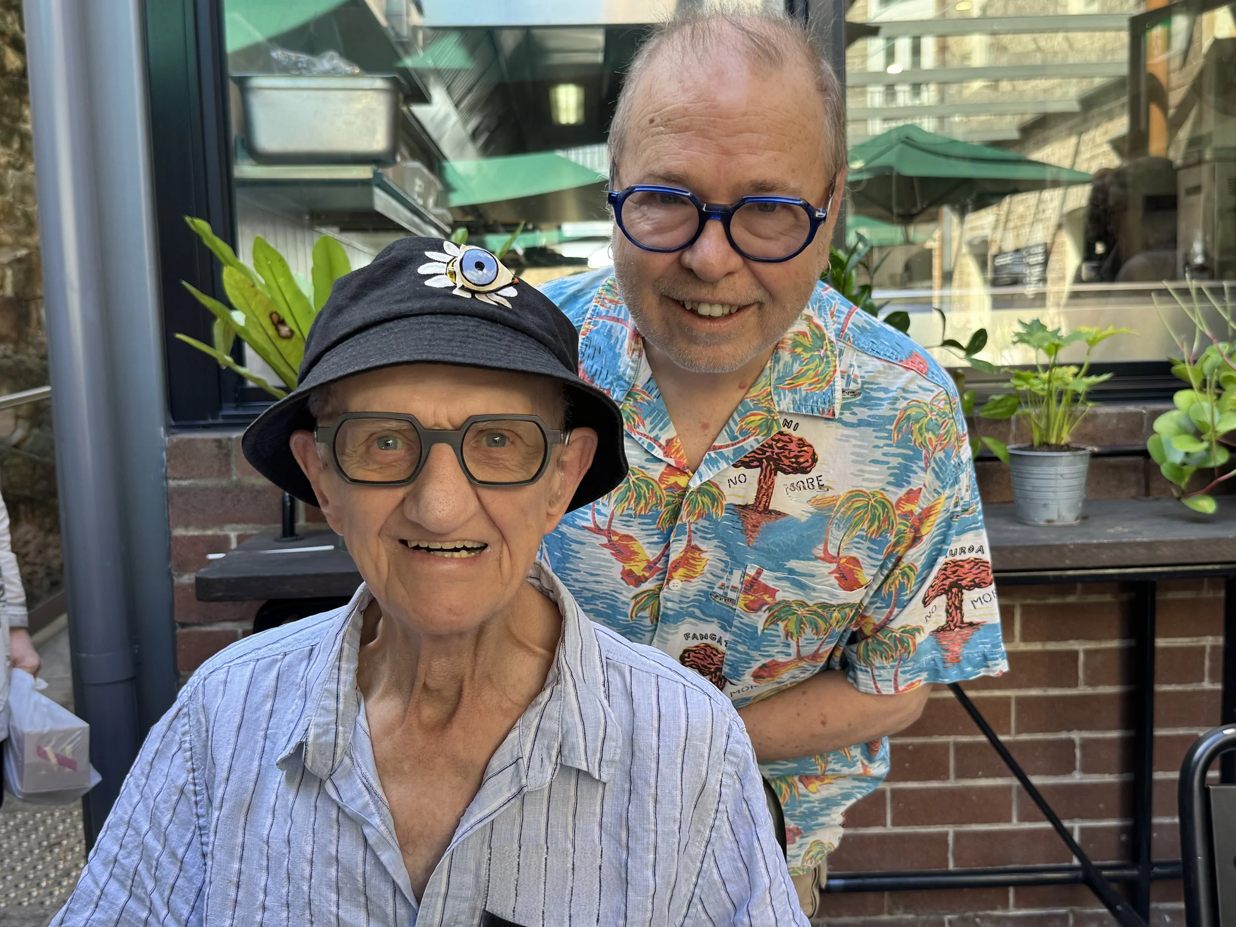 A man with blue glasses and a colorful tropical shirt standing beside a woman with glasses and a black hat with a bee decoration. They are outdoors in front of a brick wall and plants.