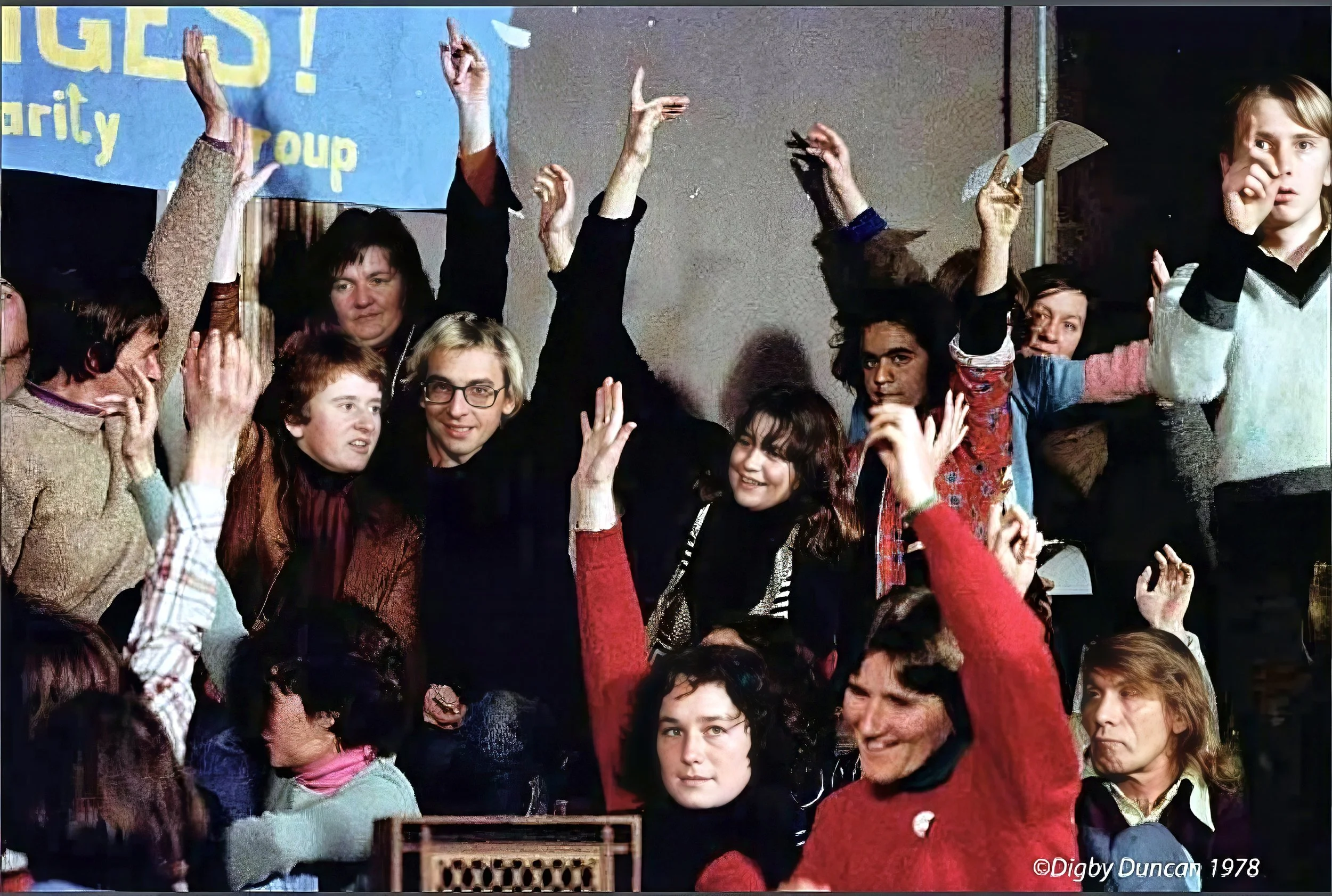 A group of people of various ages gathered closely together, some with raised hands, in what appears to be a lively indoor event or gathering.