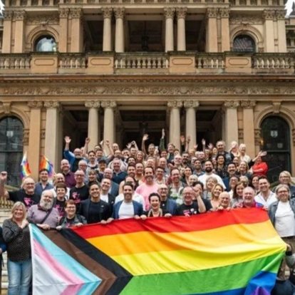Large group of people gathered in front of a historic building, holding a rainbow pride flag with a black and brown stripe, celebrating LGBTQ+ pride.
