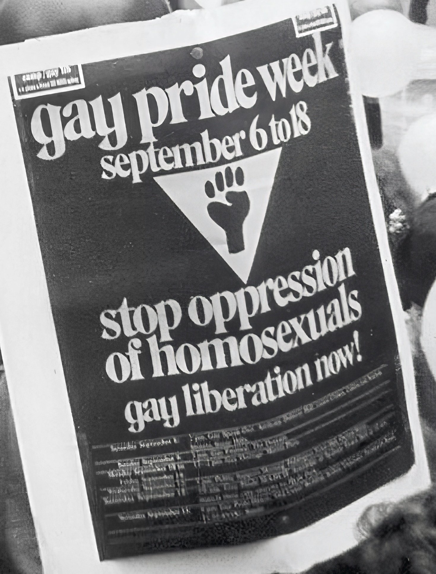 A newspaper headline in black and white reading, "Gay Pride Week September 6 to 18" with a symbol of a rainbow flag incorporating a fist. The headline also states, "Stop oppressors of homosexuals".