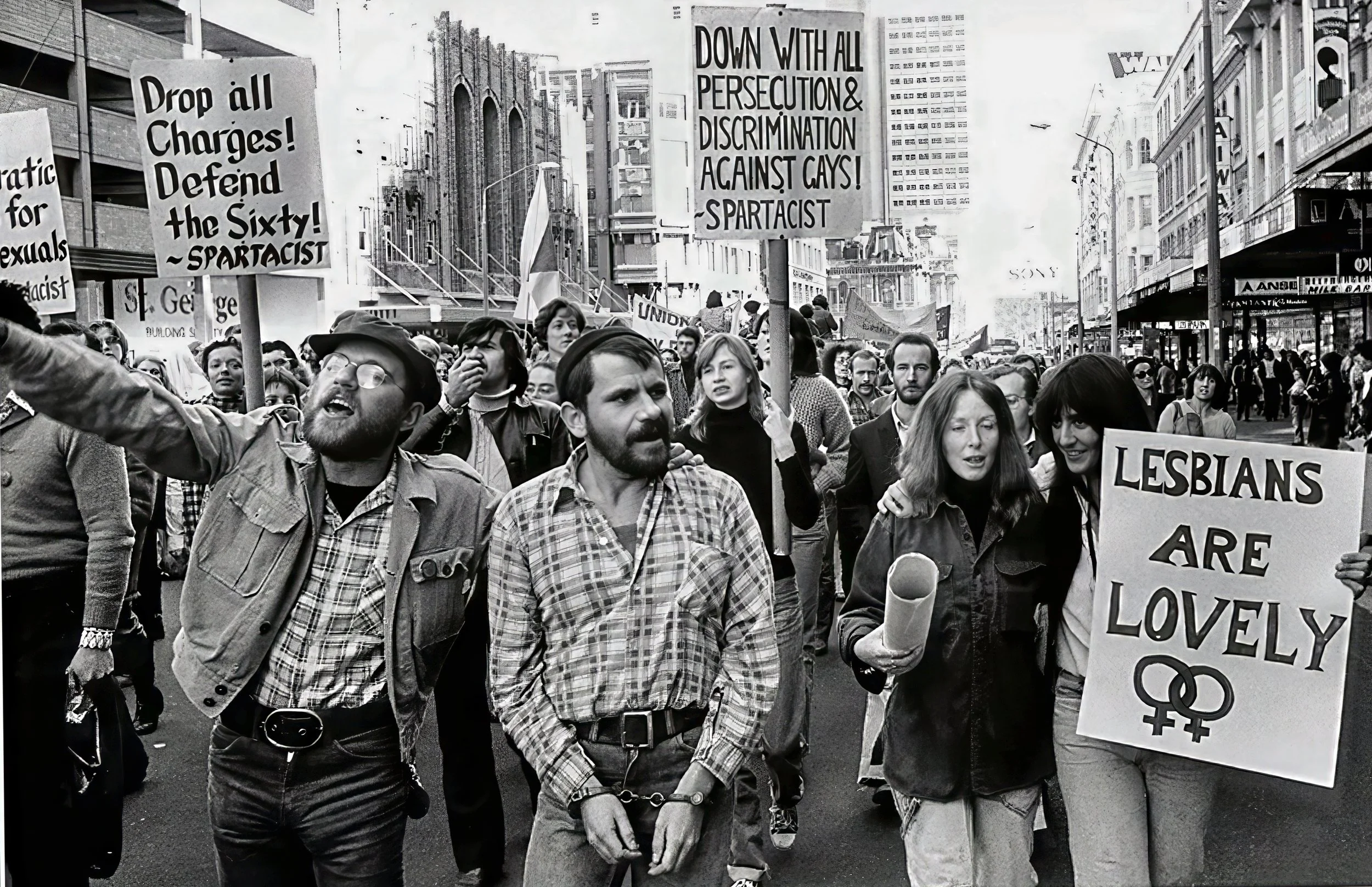 A black and white photo of a street protest with a crowd holding various signs. Signs include messages such as 'Drop all charges! Defend the sixty! - SPARTACIST' and 'Down with all persecution & discrimination against gays! SPARTACIST'. Protesters are gathered among tall city buildings with some storefronts visible.