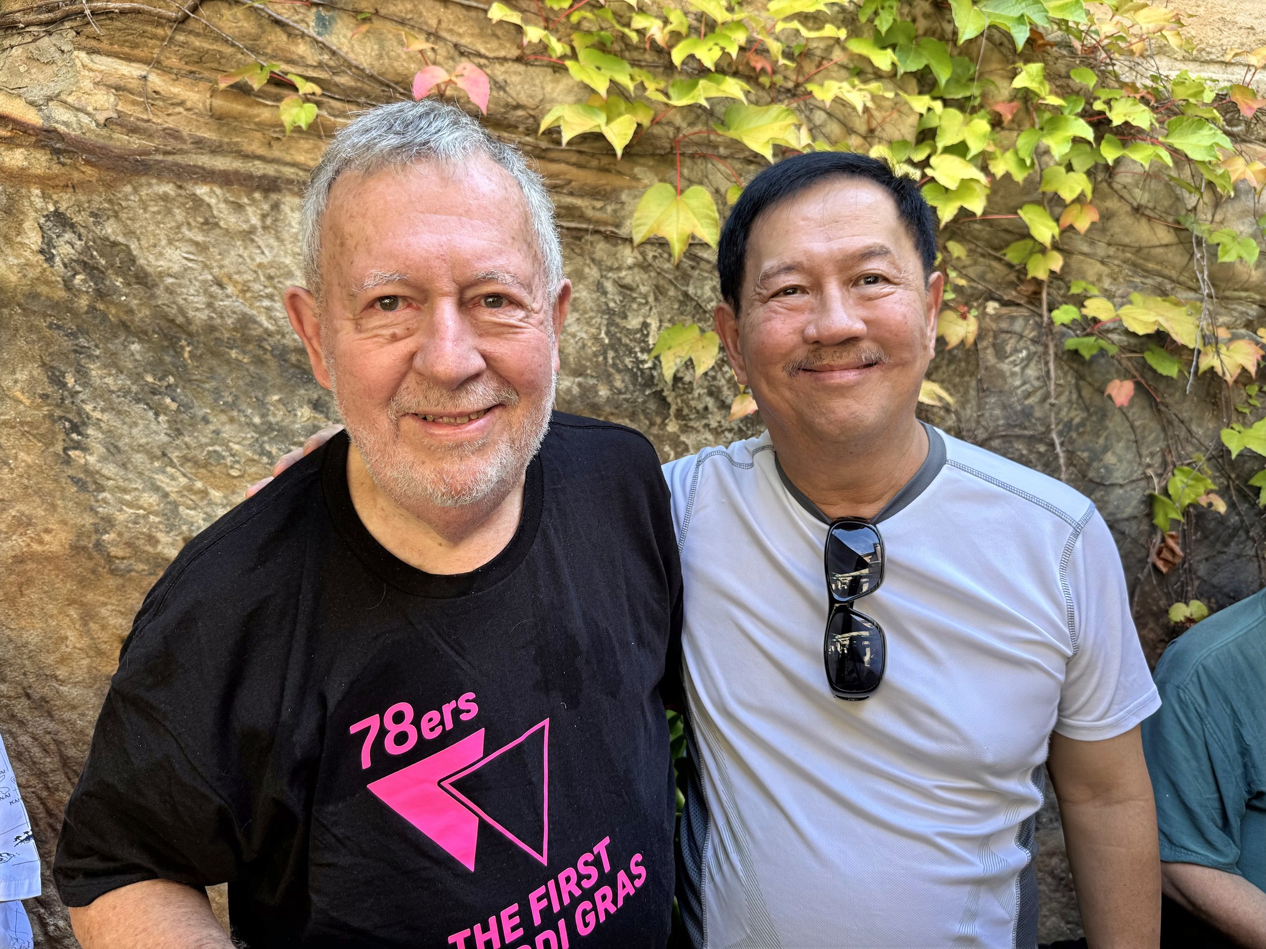 Photo of two men standing outdoors in front of a rock wall with green and yellow ivy. The man on the left has gray hair and beard, wearing a black T-shirt. The man on the right has black hair, wearing a light gray T-shirt and sunglasses hanging from his shirt collar, smiling at the camera.