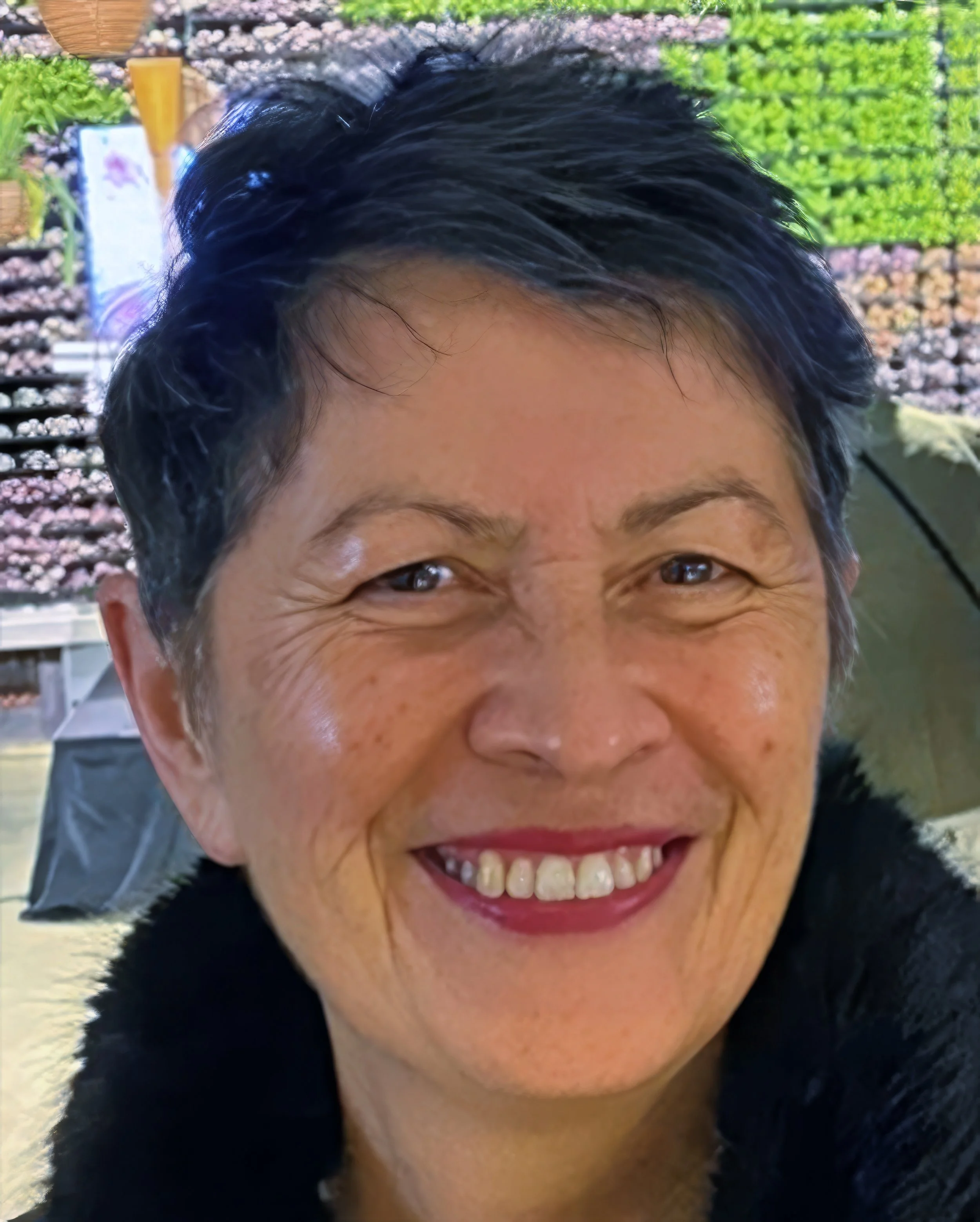 Close-up of a woman smiling outdoors with trees and a building in the background.