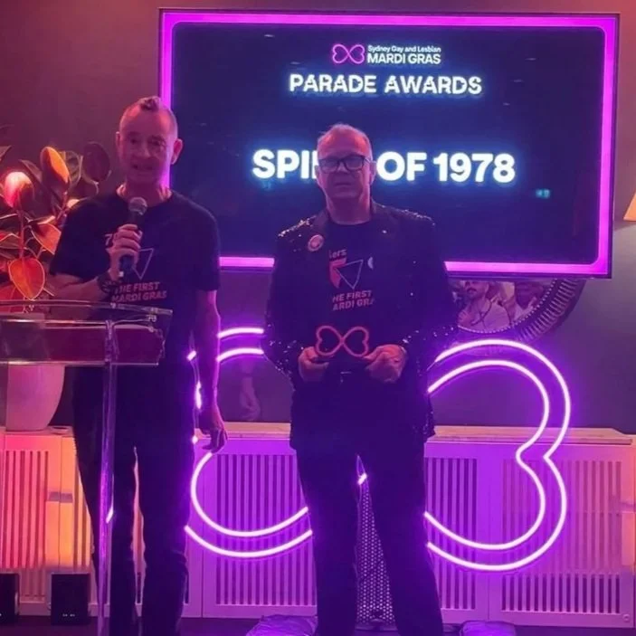 Two individuals stand on stage during the Sydney Gay and Lesbian Mardi Gras Parade Awards ceremony, with a large neon '03' sign and a screen displaying 'Parade Awards' and 'Spirit of 1978' behind them. One person is holding a microphone, and the other is holding an award.