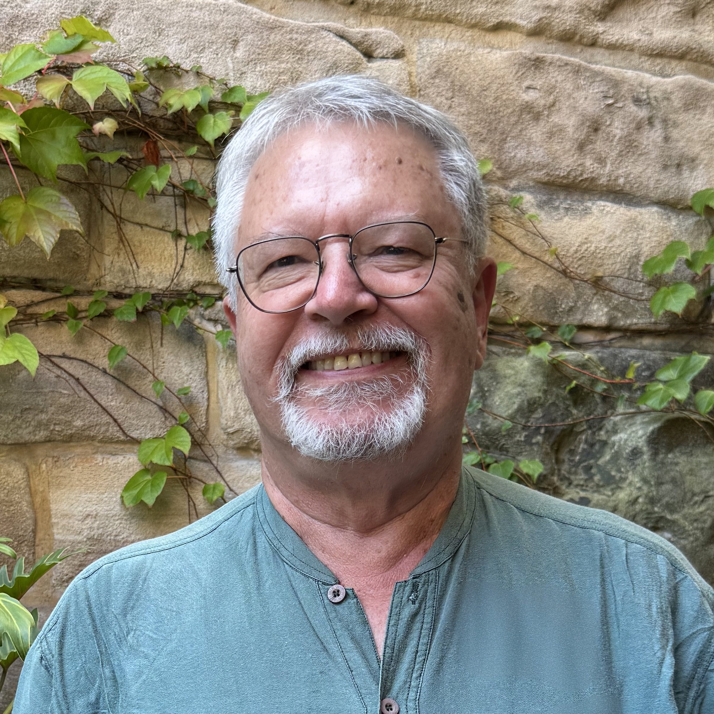 A smiling elderly man with gray hair and glasses, wearing a teal shirt, standing in front of a stone wall with climbing green ivy.
