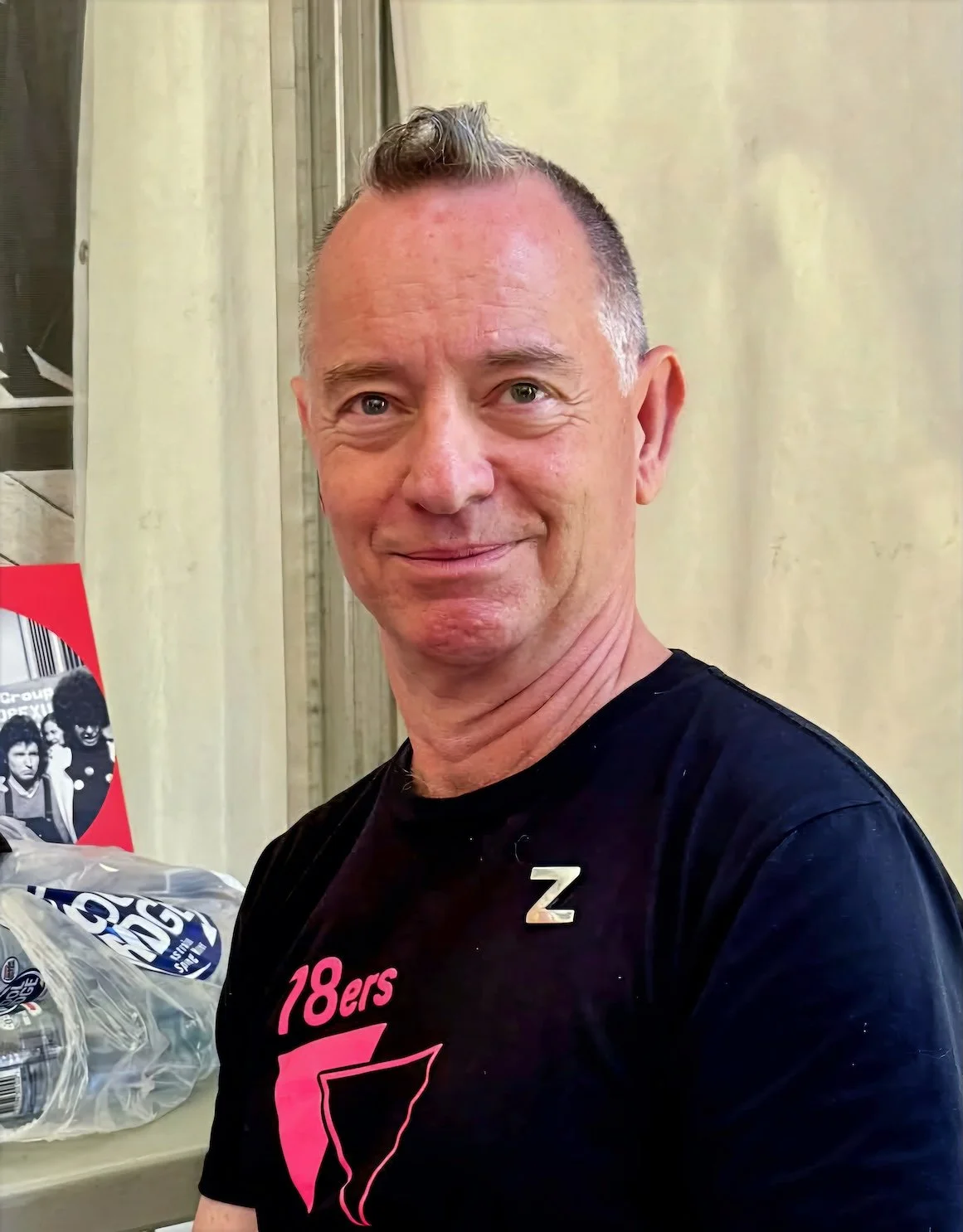 A smiling man with short brown hair and a small curl on top, wearing a black T-shirt with pink and white text and a silver pin in the shape of a letter Z, standing indoors against a beige wall.