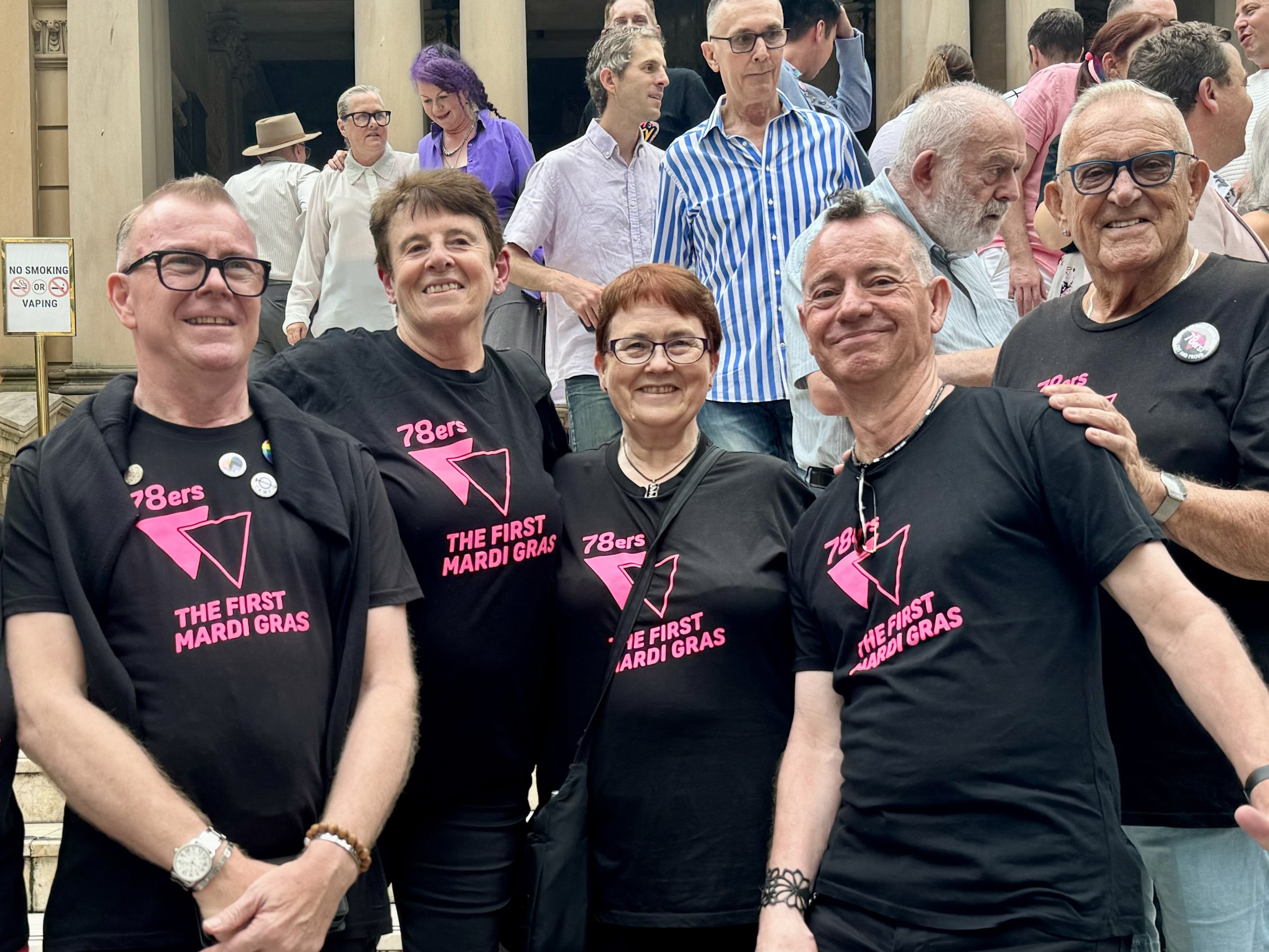 Group of people gathered for a celebration, some wearing black t-shirts that say '78ers The First Mardi Gras'. They are smiling, standing outdoors in front of a building with columns, with a mix of men and women of various ages.