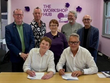 Seven people, five men and two women, gathered around a table with papers, at an event called 'The Workshop Hub' as seen on the purple sign behind them, inside a room with pink walls and purple decorations.