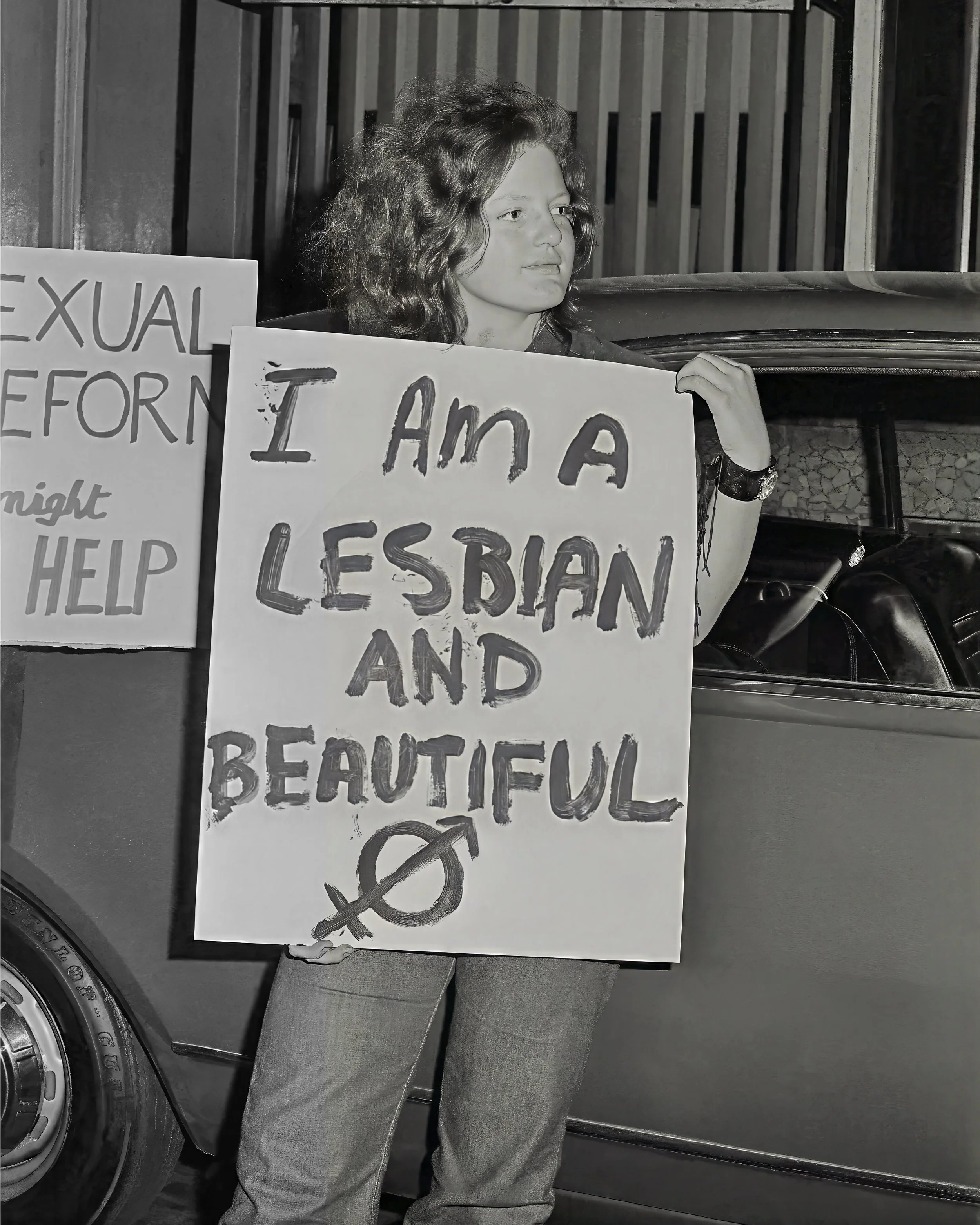 A person is holding a protest sign that reads, 'I am a lesbian,' in front of a car with a tire visible at the bottom left. The person has curly hair and is standing outdoors.