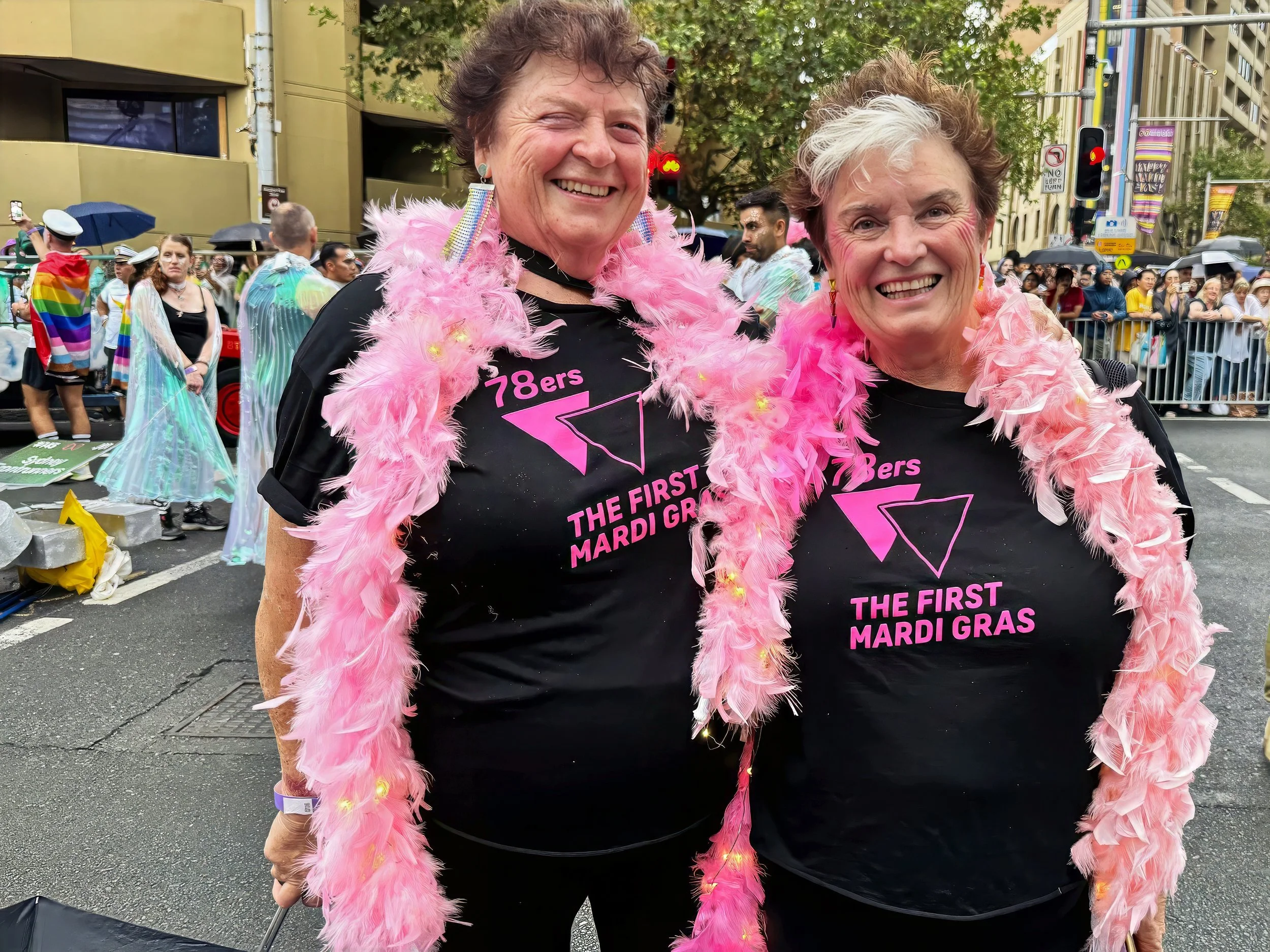 Two women smiling at a Pride parade, wearing pink feather boas and black T-shirts with pink text celebrating the first Mardi Gras, in a city street with parade floats, people in rainbow costumes, and spectators in the background.