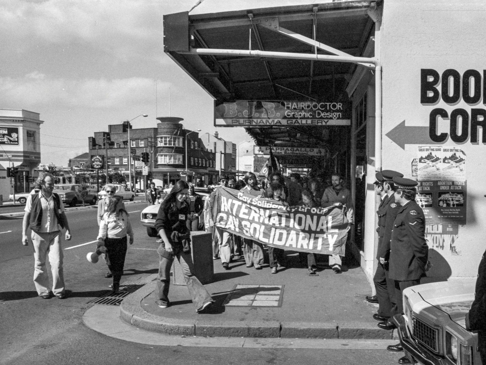 A group of protesters holding a banner that reads 'International Gay Solidarity' march on a city sidewalk. Some police officers stand nearby, and pedestrians walk past.