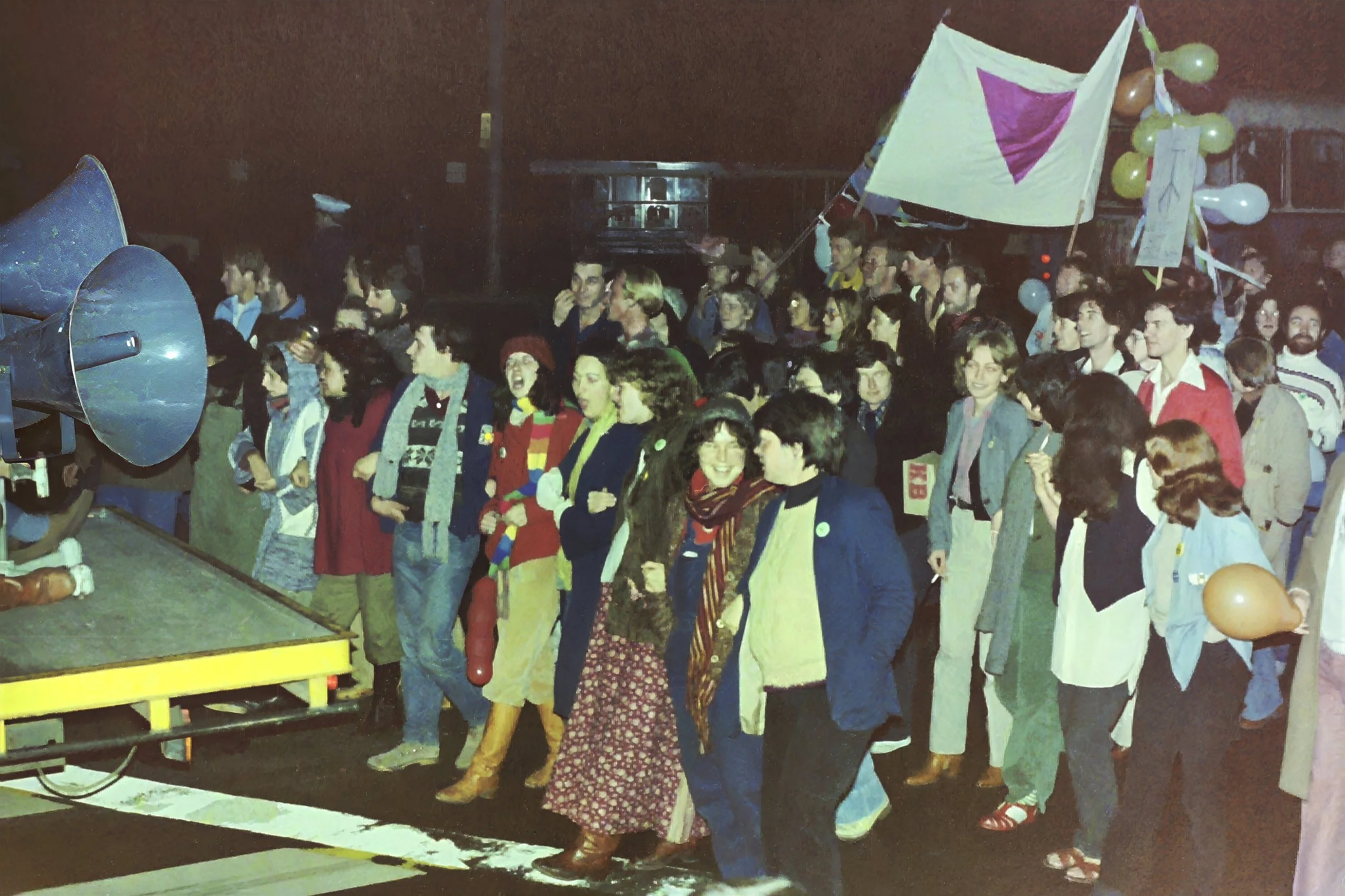 A group of people gathered at a nighttime outdoor event, some young adults, some older, standing in line or waiting, with a speaker and balloons decorated for celebration.