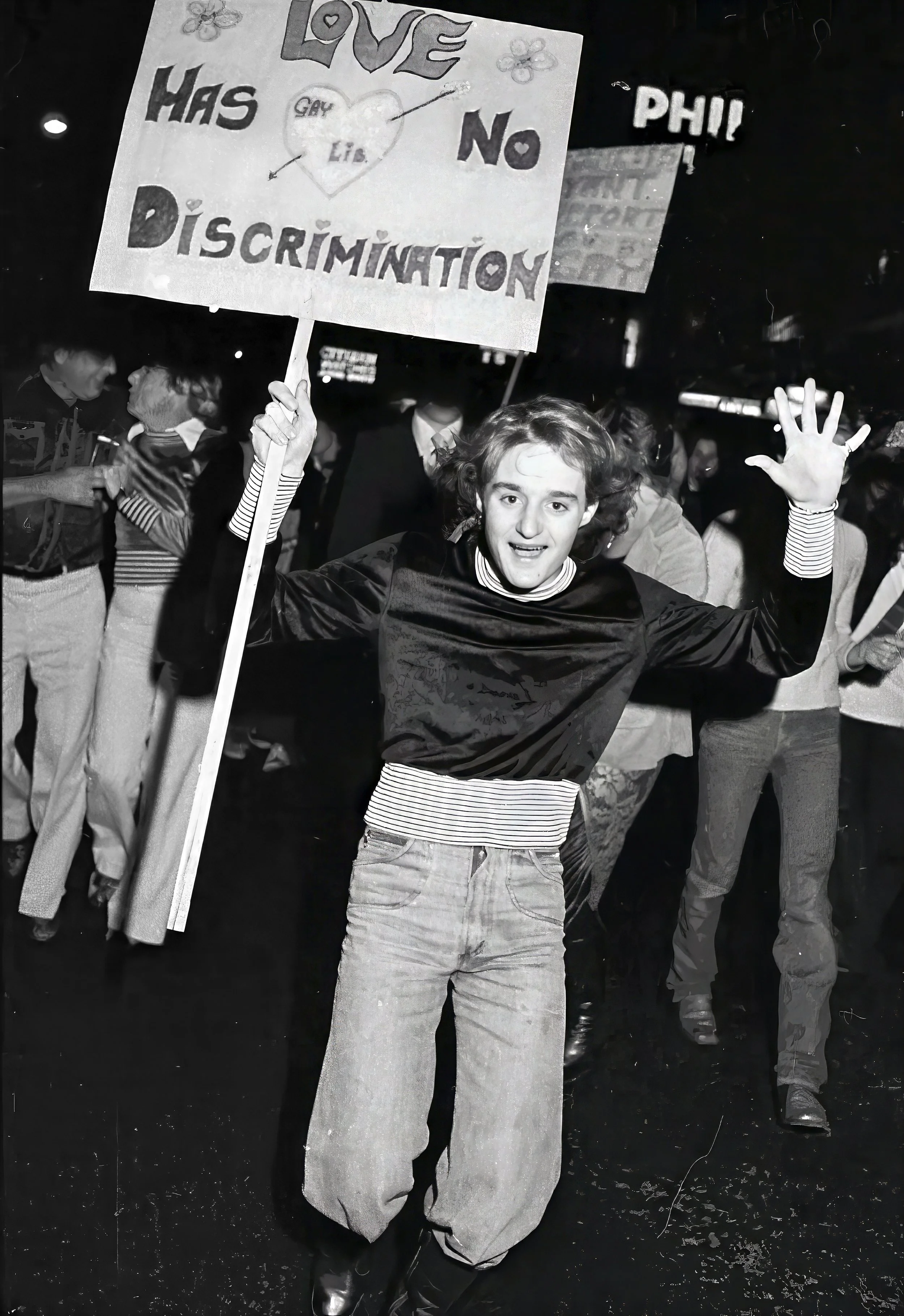 A young man at a protest holding a sign that reads: 'Love has no discrimination' with a heart and butterflies. He is smiling and waving at the camera, with other protesters in the background.