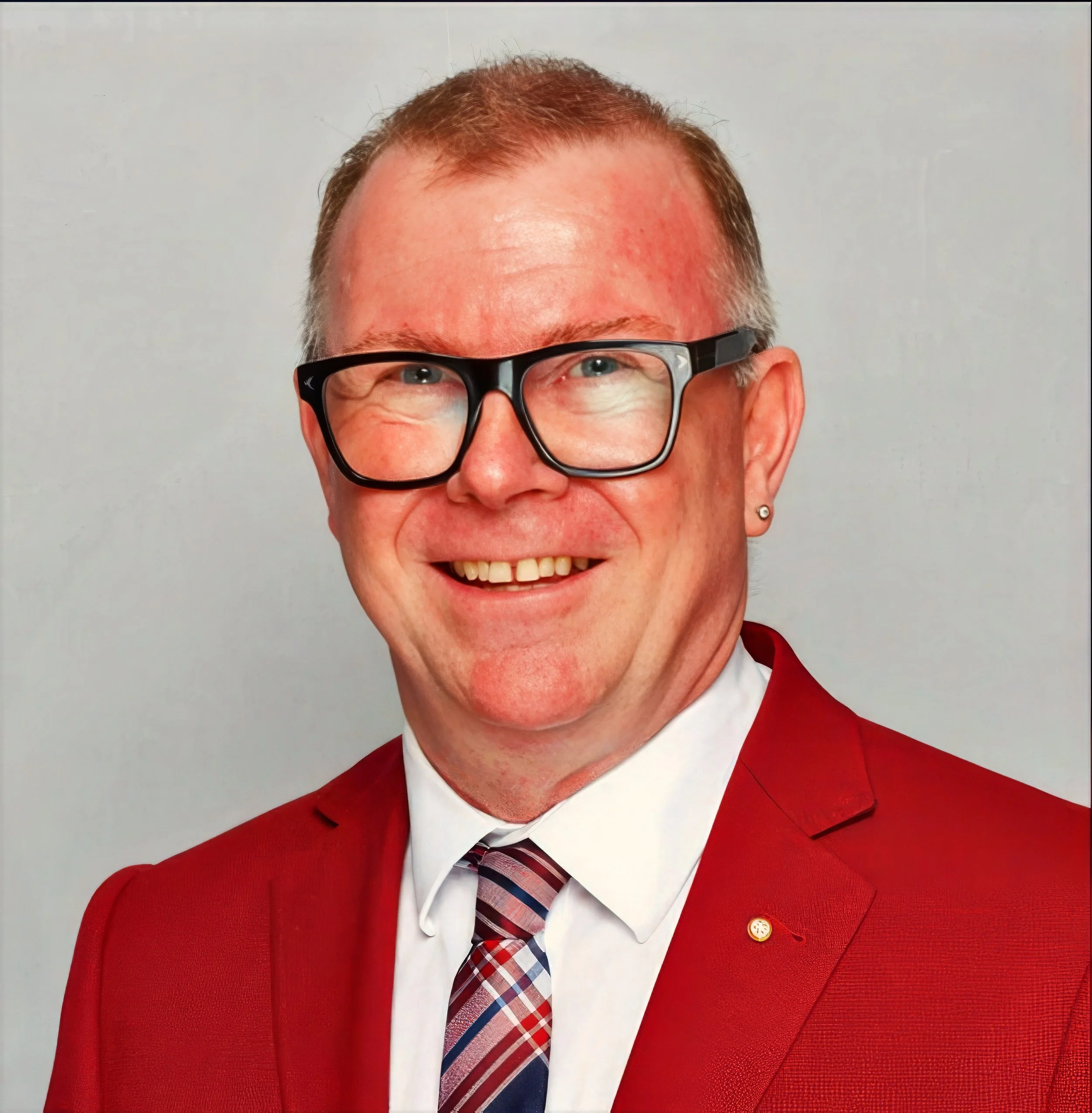 A man with glasses, short gray hair, and a small earring in his left ear, wearing a red blazer, white shirt, and a plaid tie.