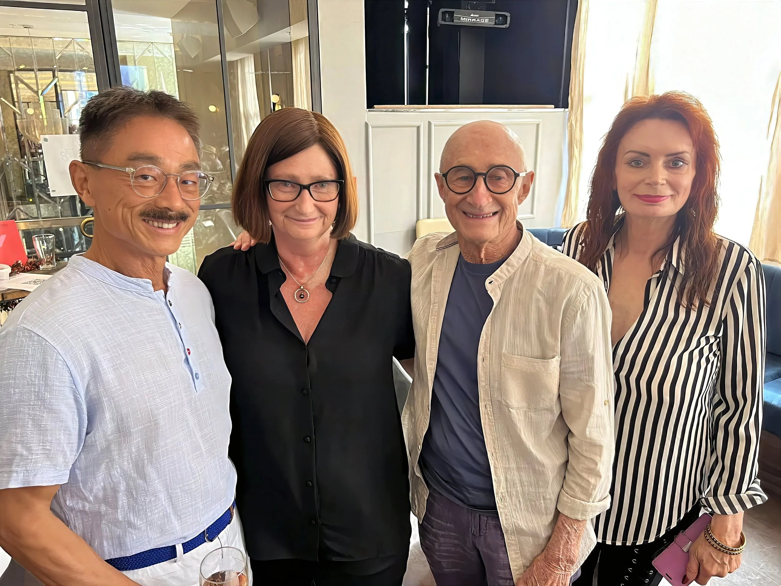 Four people standing together indoors, smiling, with a glass window and curtains in the background.