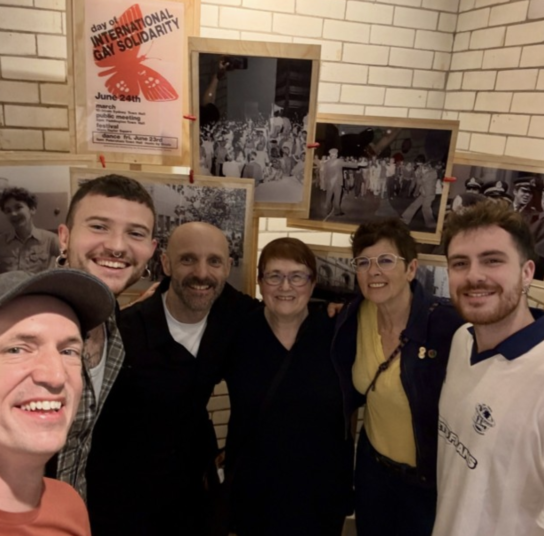 Group of six people smiling for a selfie in front of black-and-white photographs and a poster about LGBTQ+ pride on a brick wall.