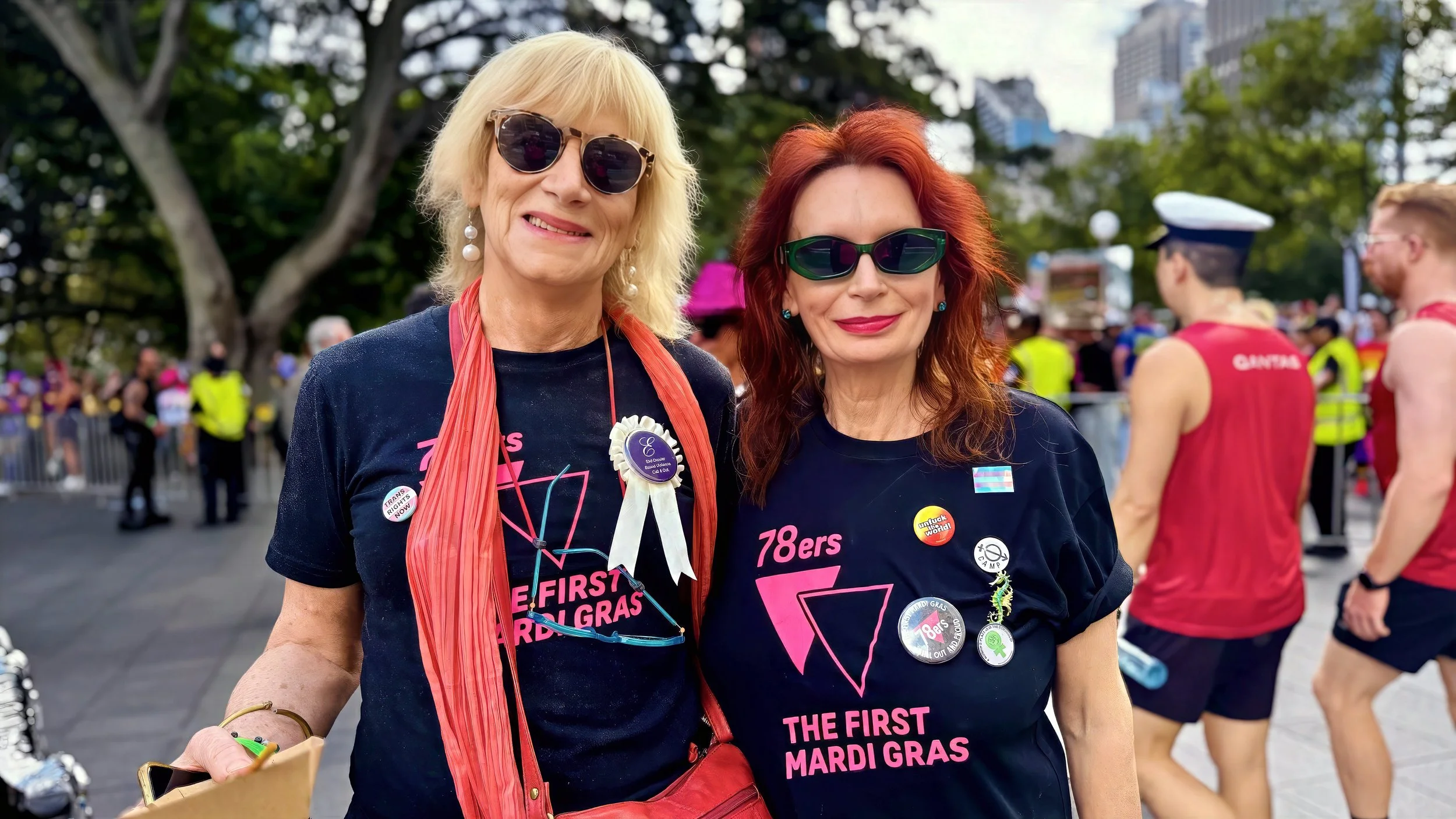 Two women with sunglasses and Mardi Gras buttons standing together at a parade, with people and trees in the background.