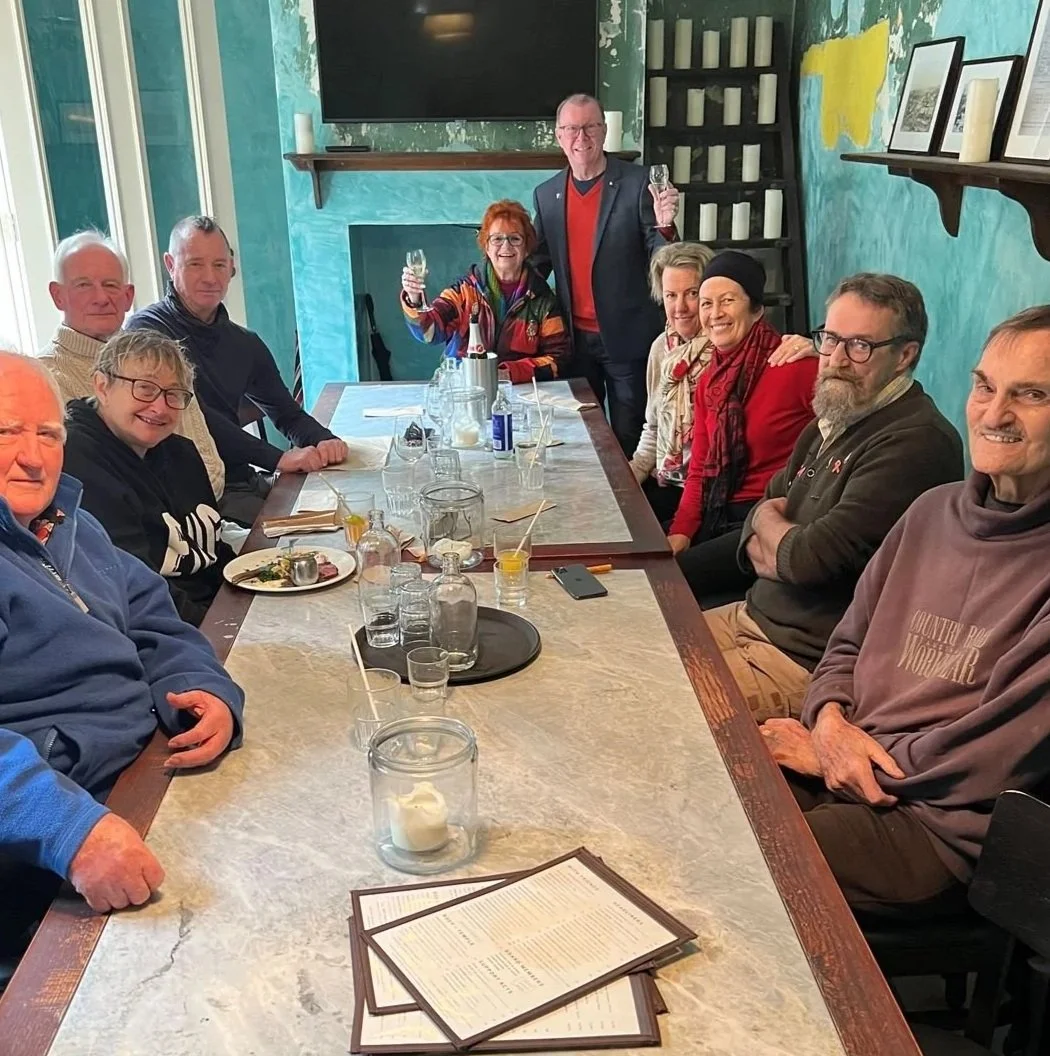 A group of twelve people gathered around a long dining table in a restaurant, celebrating with two individuals standing and holding wine glasses, smiling. The table has plates, glasses, and a menu. The background features teal-colored walls with framed pictures and a large TV screen.