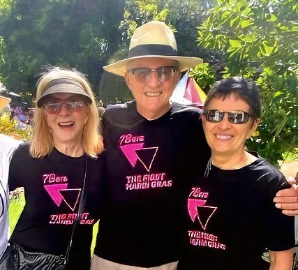 Three people standing outdoors, smiling, wearing black shirts with pink text and graphics, and sunglasses, with trees and a pink umbrella in the background.
