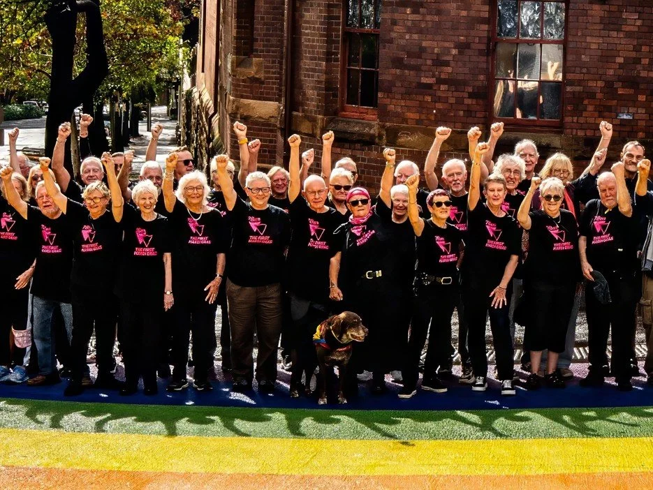 A diverse group of 78ers participate in an outdoor rally or march, wearing matching black T-shirts with pink graphics and lettering, raising their fists in solidarity behind a large rainbow-colored banner.