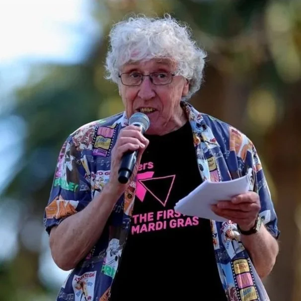 An older man with curly white hair and glasses speaking into a microphone while holding a paper, wearing a colorful patterned shirt and a black T-shirt with pink text.