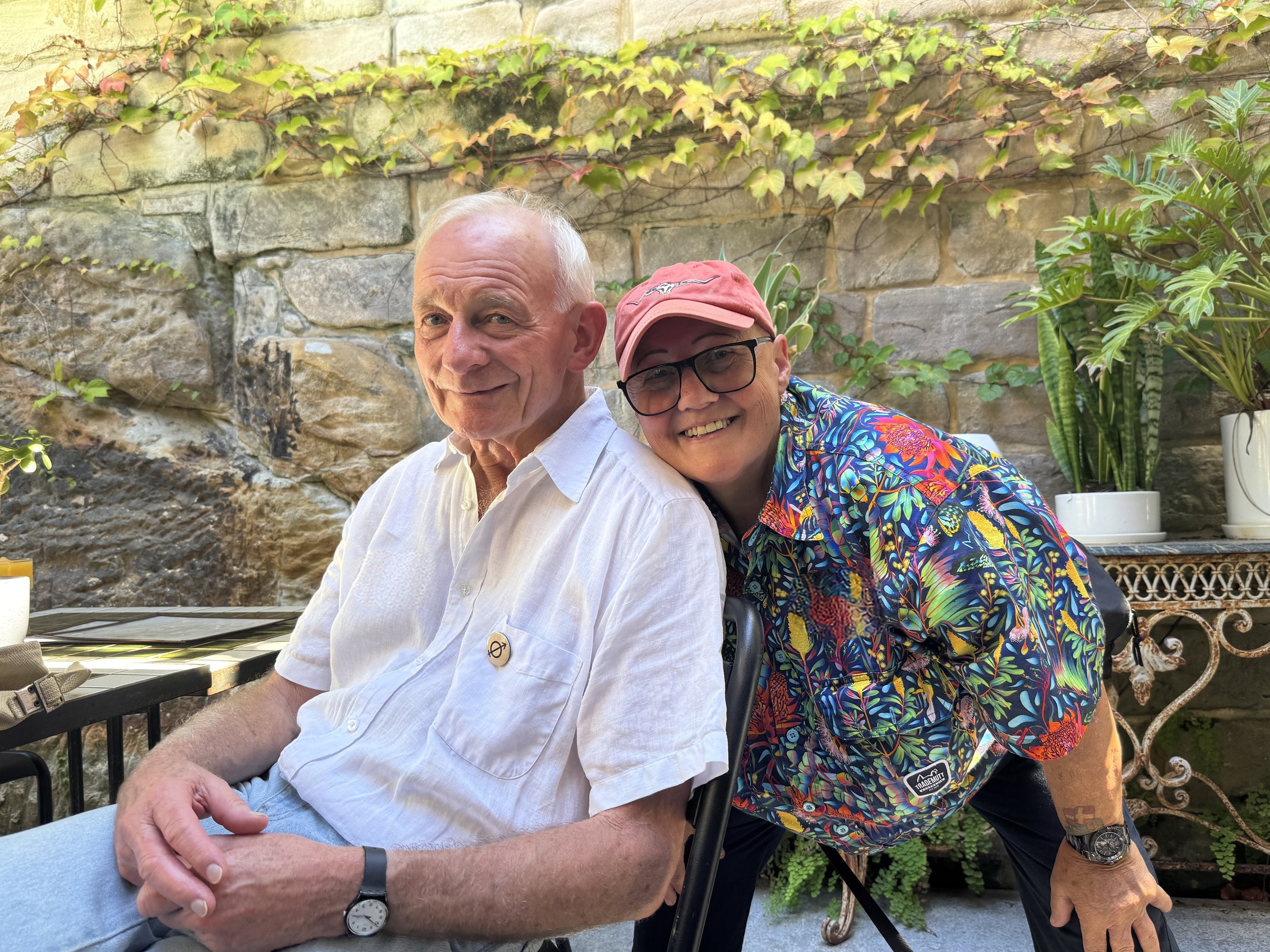 A smiling older woman wearing glasses, a pink cap, and a colorful floral shirt leaning over a seated elderly man with short white hair, wearing a white dress shirt, in an outdoor setting with a stone wall and potted plants in the background.