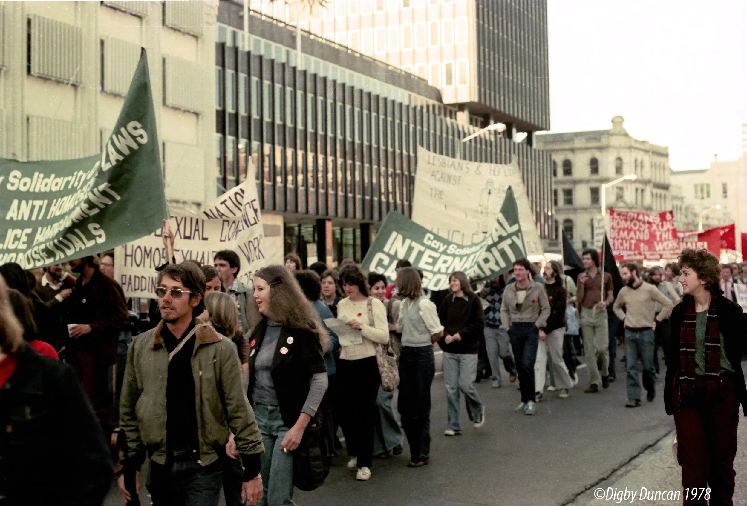 A group of people participating in a protest or march on a city street with multiple banners and signs advocating for LGBTQ+ rights, with tall buildings in the background.