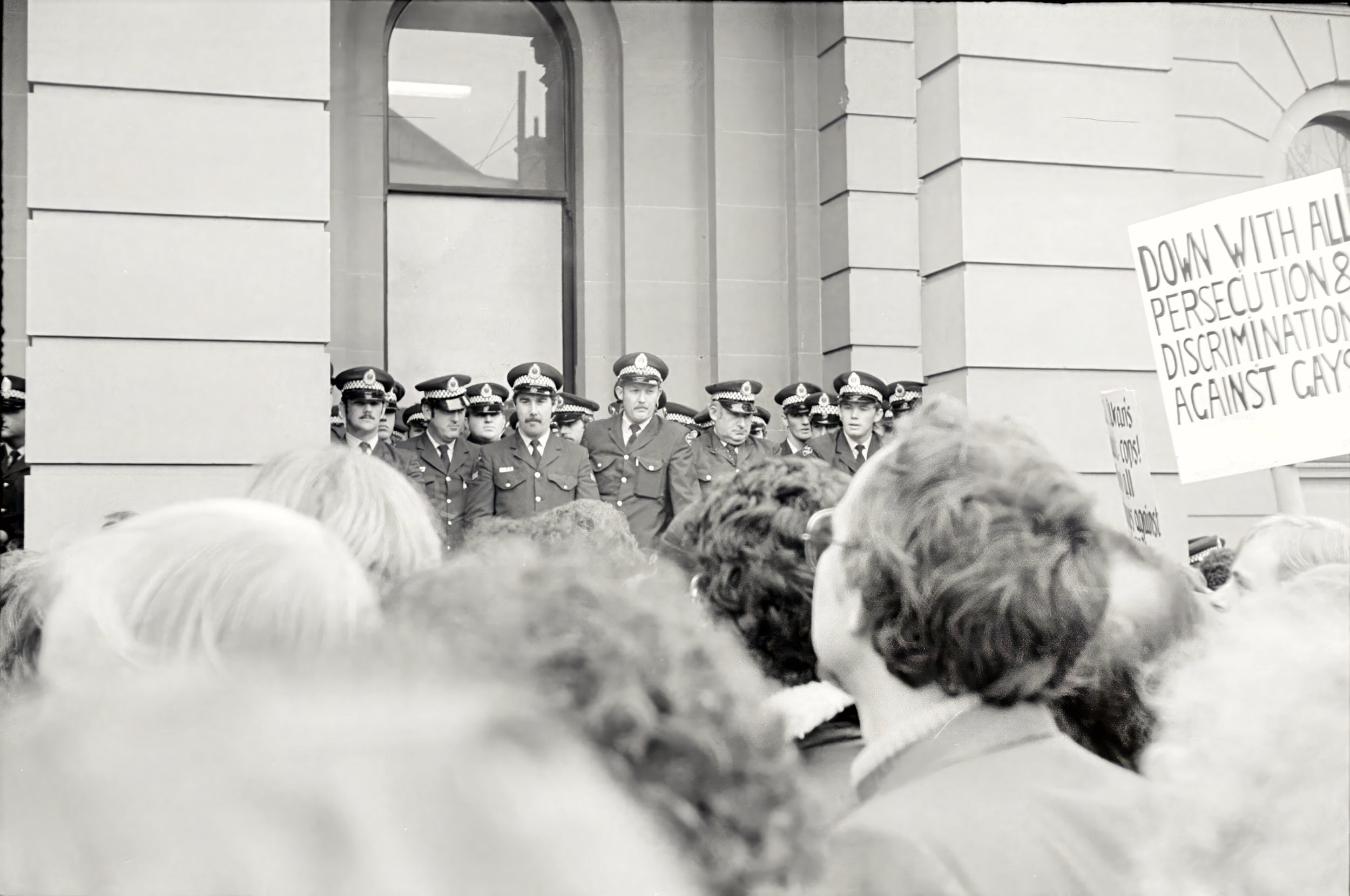 A group of police officers in uniform standing in front of a building with large windows during a protest or demonstration. A sign held by a protester in the foreground reads, "Down with all persecution & discrimination against gays."