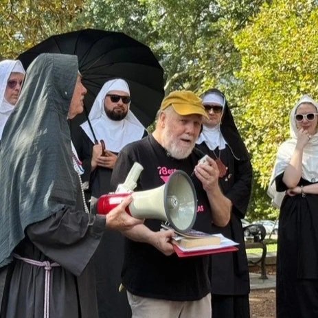 A man speaking into a megaphone outdoors, surrounded by nuns in black and white habit uniforms, some with umbrellas and sunglasses.
