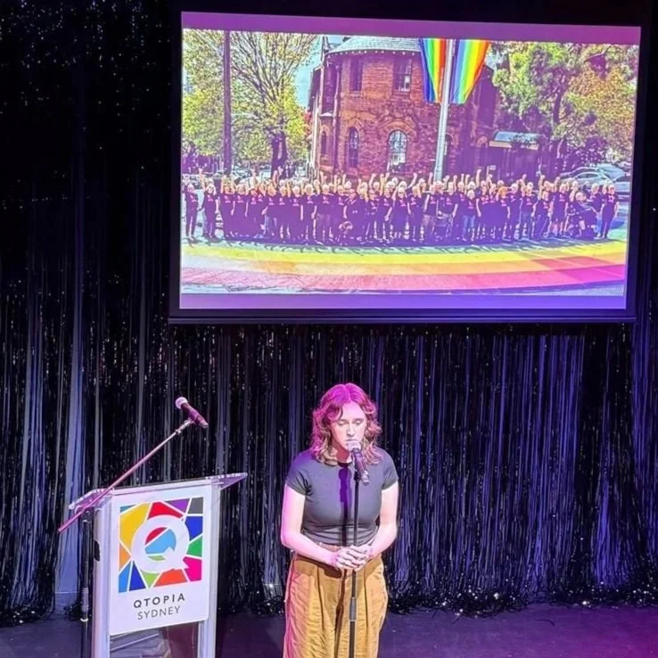 A woman with wavy brown hair wearing a black shirt and yellow pants standing on stage in front of a microphone. Behind her is a black glittering curtain and a podium with a colorful logo and the words "Qtopia Sydney." A large screen above shows a group photo of many people lined up outside in front of a brick building with trees, some rainbow flags, and a rainbow crosswalk.