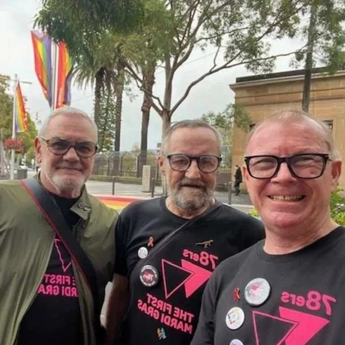 Three men standing outdoors, wearing black T-shirts with pink text and buttons supporting breast cancer awareness, with rainbow flags and trees in the background.