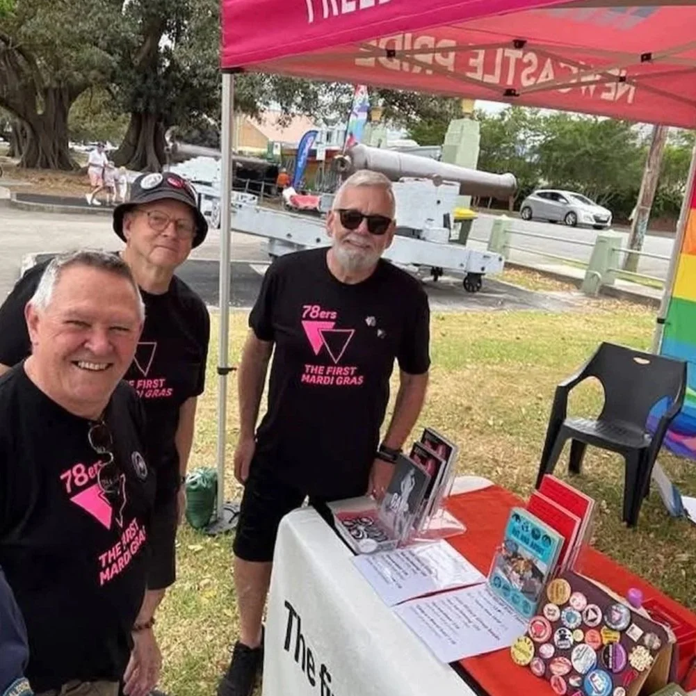 Three men smiling at a table under a pink canopy tent at an outdoor event during Mardi Gras. The men are wearing black T-shirts with pink and white lettering that says '78ers THE FIRST MARDI GRAS.' The table is covered with various flyers, buttons, and pamphlets, with a rainbow-colored banner visible to the right.