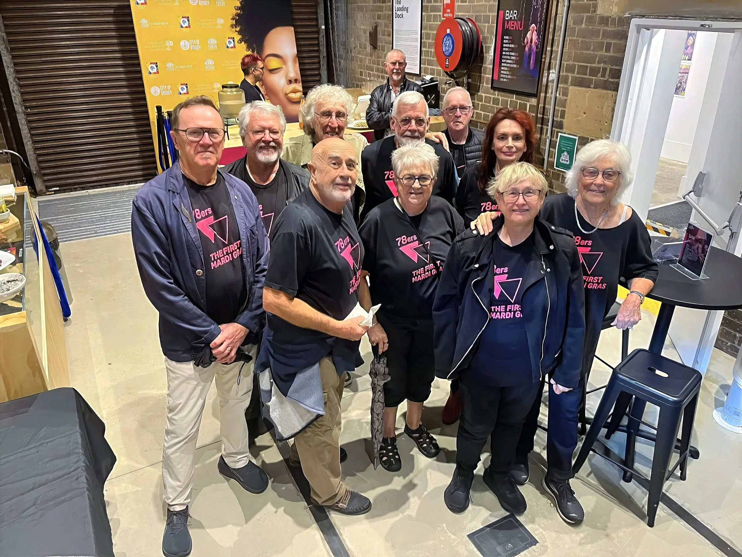 Group of people at an indoor event, wearing black shirts with pink text, posing for a photo. Behind them, posters and a bar menu are visible on brick walls.