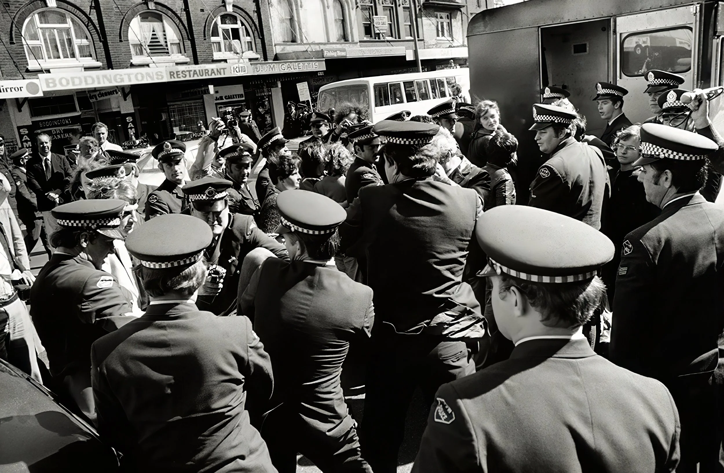 A group of police officers and civilians gathered in a street with buildings and storefronts, some wearing uniforms and hats, engaged in a discussion or confrontation in black and white.
