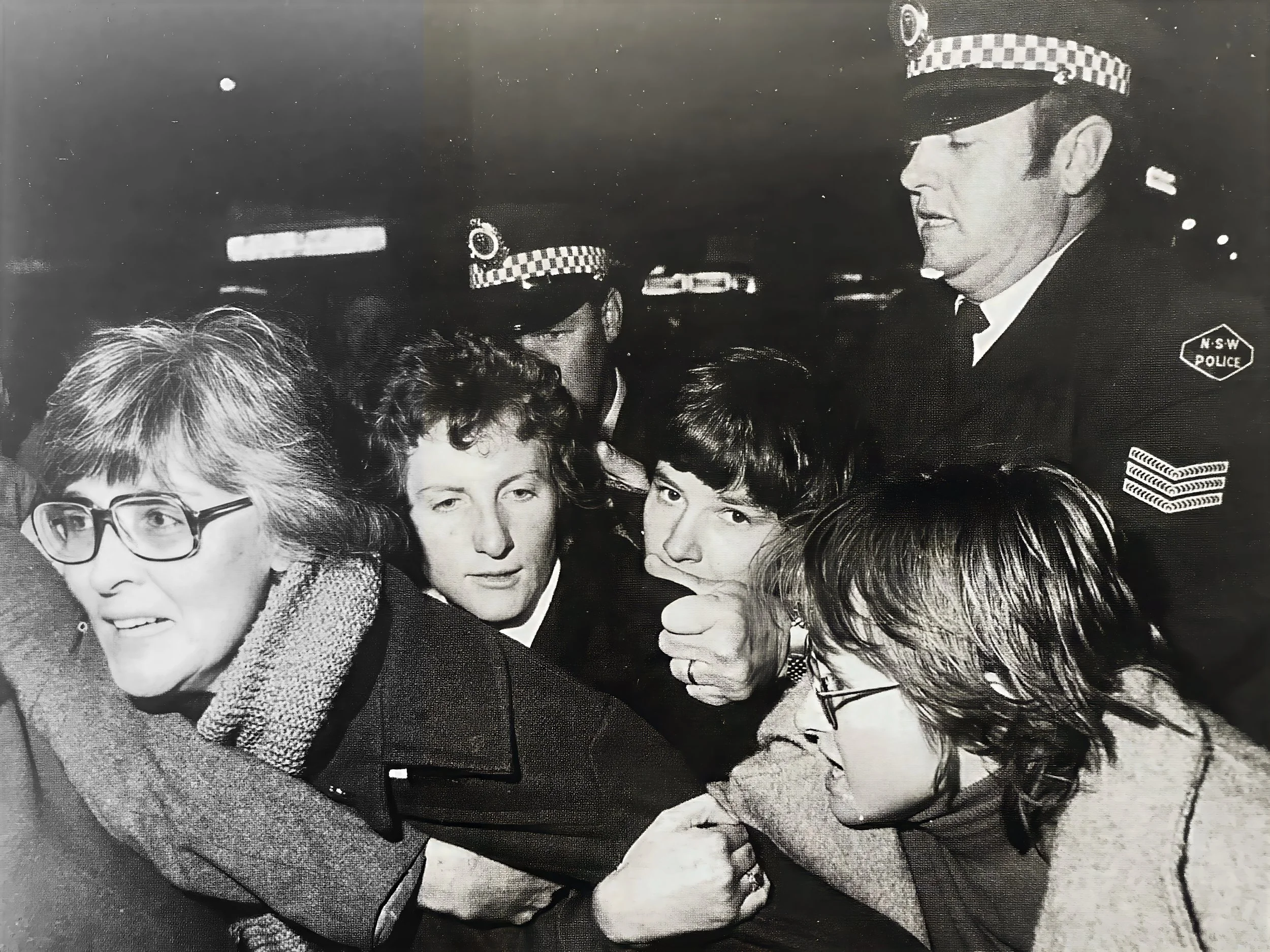 Black and white photo of five women and one police officer, all dressed in formal attire, standing close together. The police officer is wearing a uniform and hat with a checkered band, and a badge that reads 'N.S.W. POLICE'. They appear to be engaged in conversation or looking at something in front of them.