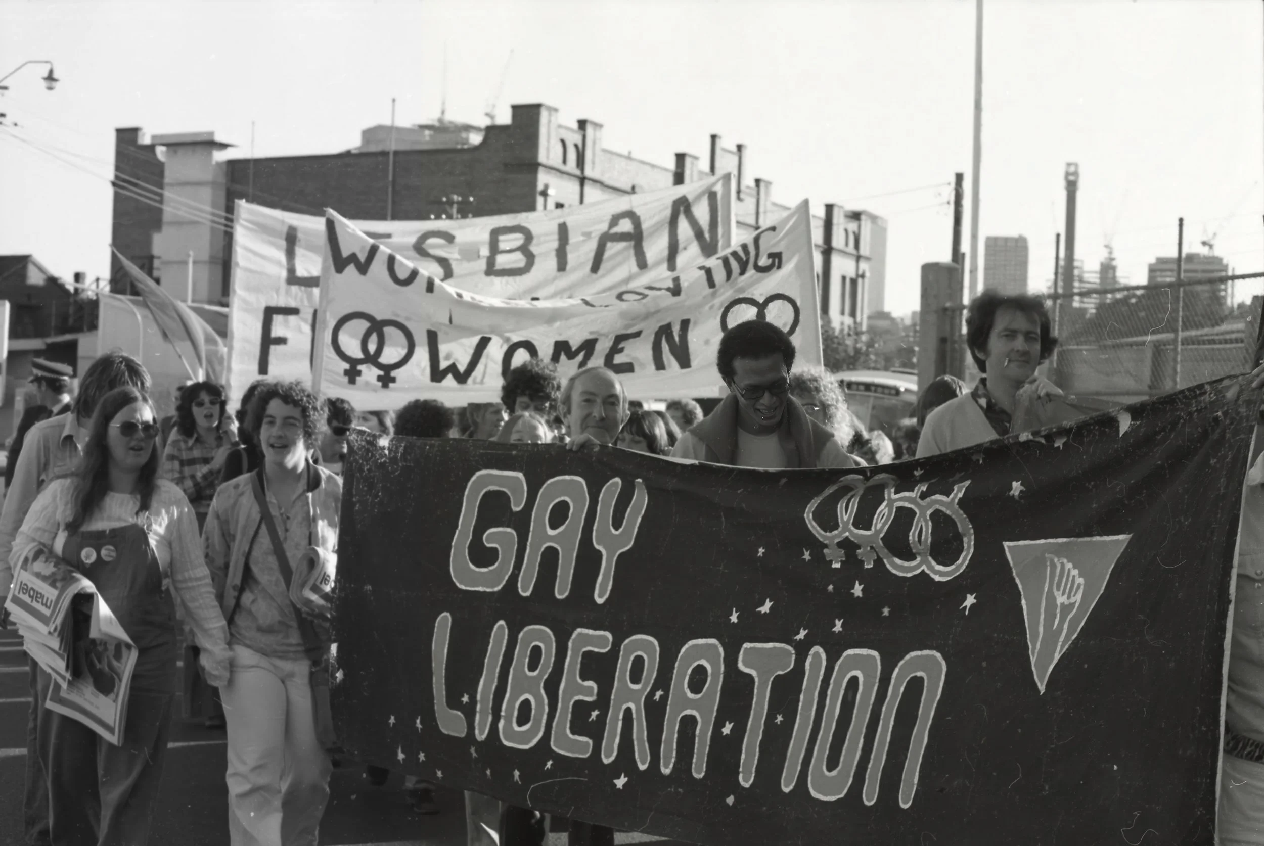 A black-and-white photo of a pride parade with people holding signs and banners. One prominent banner reads 'Lesbian Women' with symbols for female and lesbian. The crowd appears enthusiastic, with some wearing sunglasses and carrying flyers. The scene is set in an urban environment with buildings and cityscape in the background.