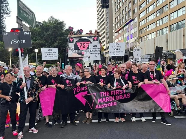 Group of people participating in a pride parade, holding a large banner that reads '78ers Mardi Gras Australia'. Several signs and banners with historical information about the LGBTQ+ community are visible in the background.