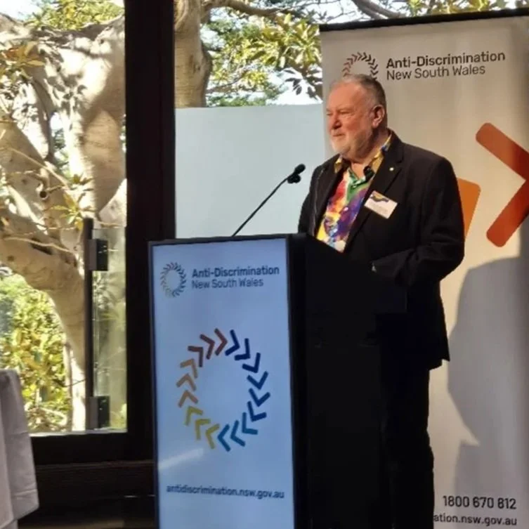 A man in a black suit and colorful shirt speaking at a podium during an event for Anti-Discrimination New South Wales, with a banner and a window showing trees in the background.
