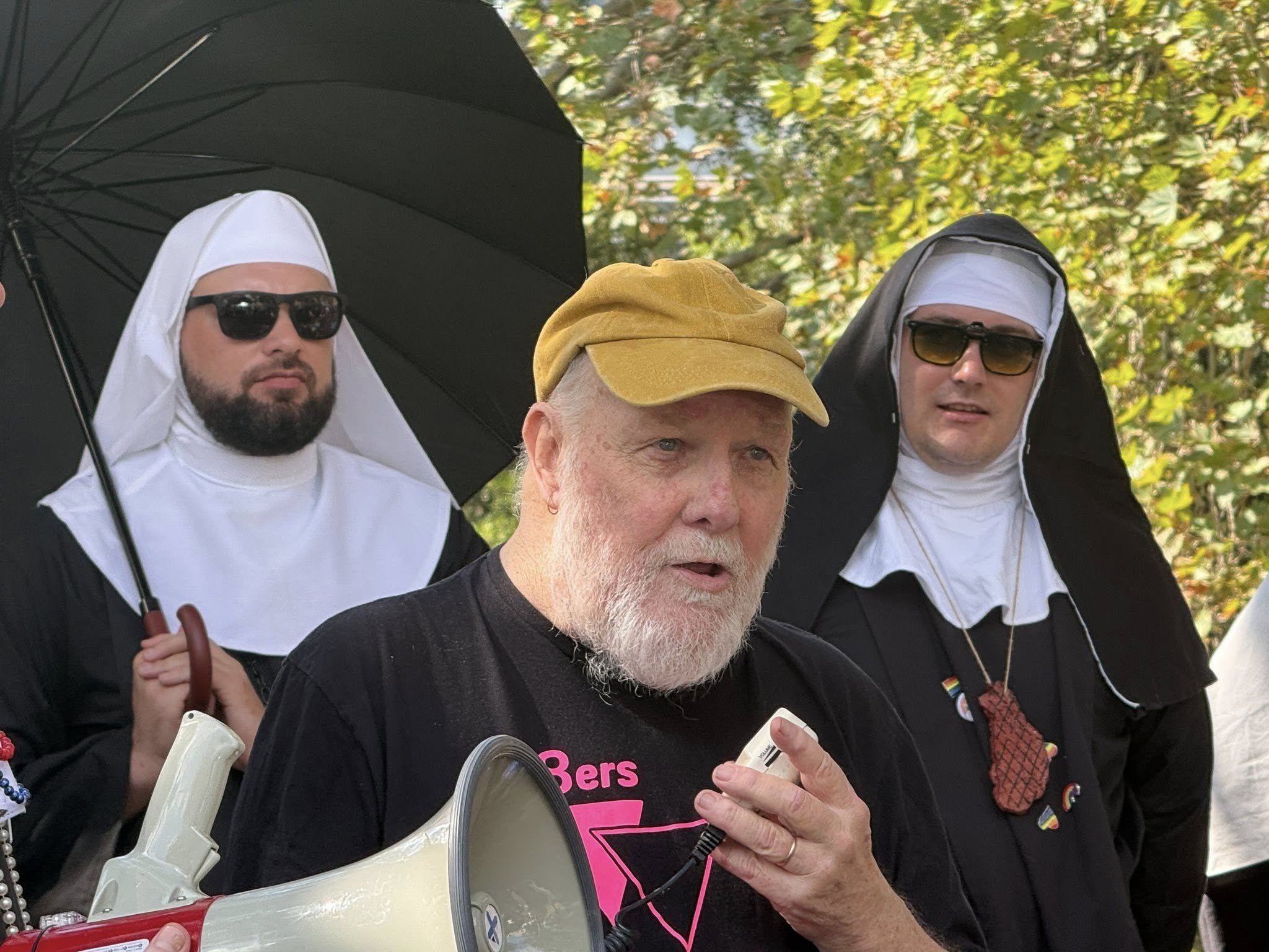 Three individuals dressed as nuns and one person wearing a tan cap and black shirt, gathered outdoors with trees in the background. The person in the foreground is holding a megaphone and appears to be speaking. The two nuns are wearing black and white habits with sunglasses. One of the nuns is holding an umbrella, and the other has an LGBT rainbow pin on her habit. The scene seems to be at a public event or gathering.