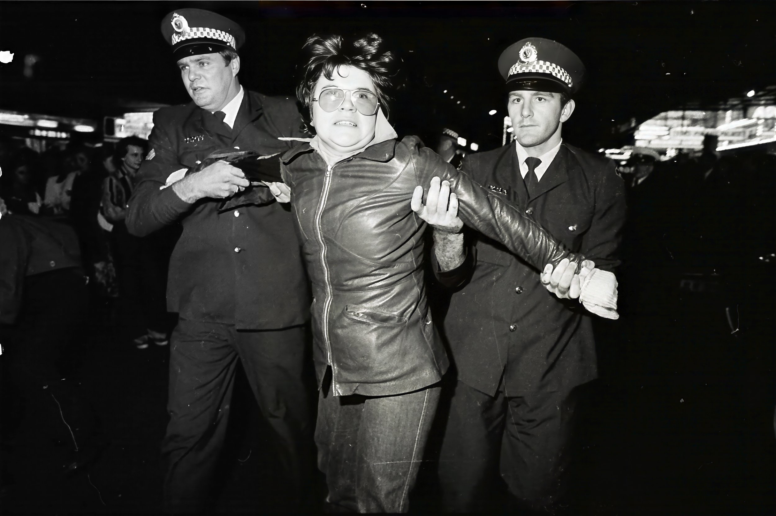 A black-and-white photograph showing a woman with glasses being detained by two police officers in uniform. The woman has short hair and is wearing a leather jacket. The setting appears to be an indoor or covered area during nighttime, with other people in the background.
