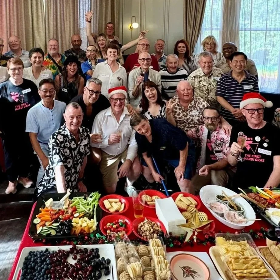 A group of people celebrating at a holiday party around a table with snacks, drinks, and festive decorations, some wearing Santa hats.