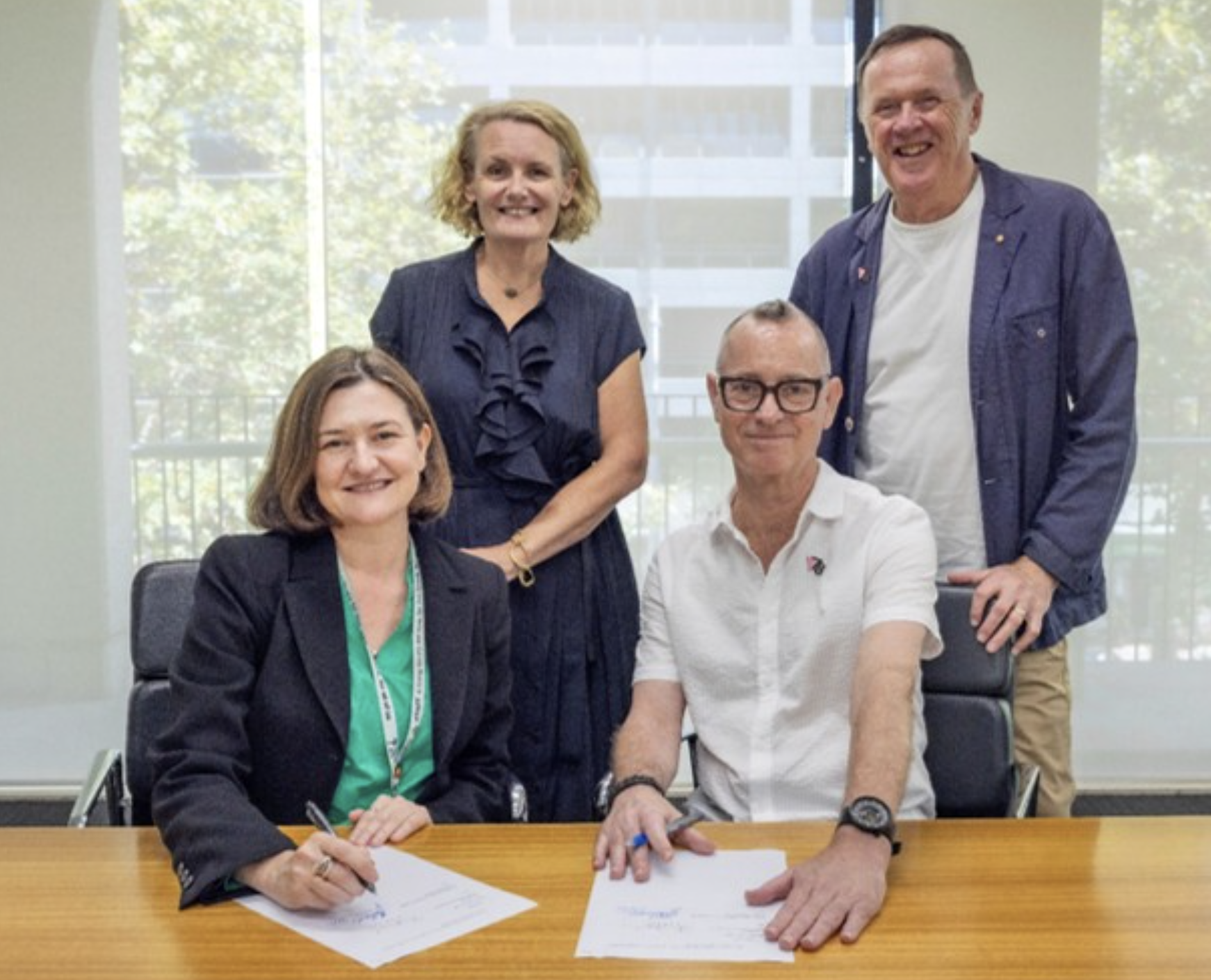 Four professionals in a meeting room, two sitting at a table signing documents and two standing behind them smiling.
