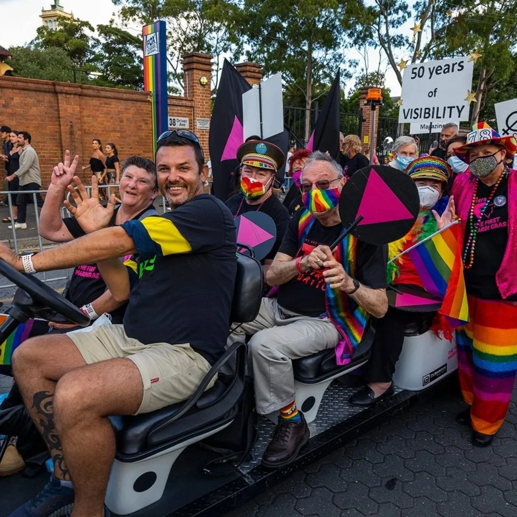 Group of people riding a parade vehicle during a Pride parade, wearing rainbow-themed masks and clothing, holding rainbow flags and pink and black flags, with a sign reading '50 years of visibility' in the background, celebrating LGBTQ+ pride.