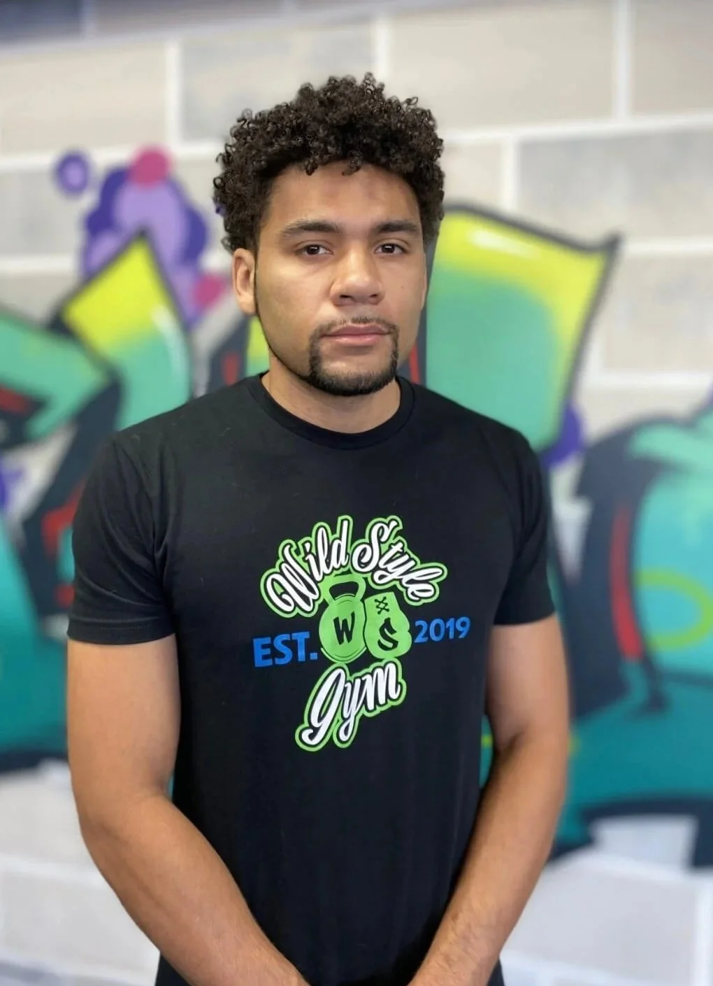 A young man with curly hair and a goatee standing in front of a colorful graffiti-covered wall, wearing a black t-shirt with the words 'World Style Gym' and a logo for a gym establishment.