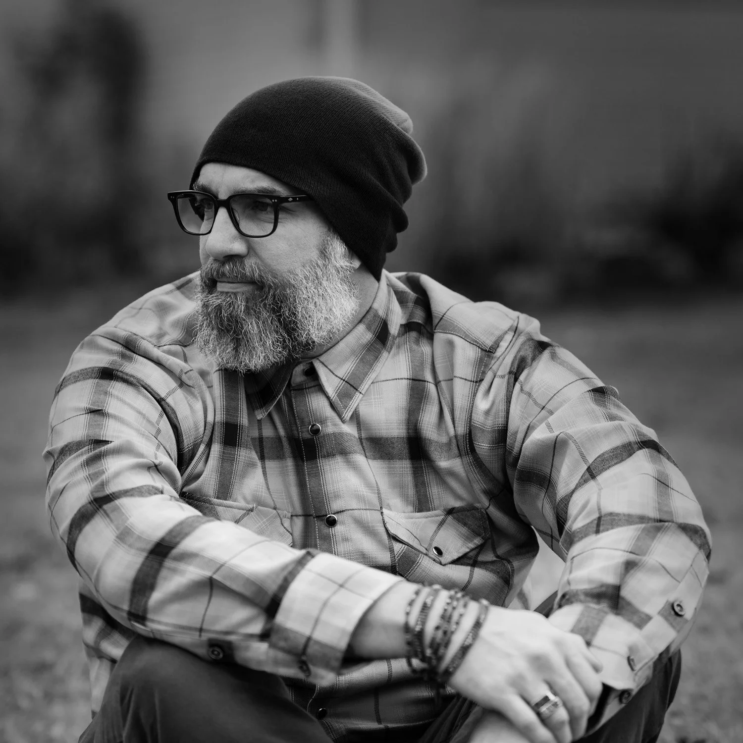 A black and white photograph of a bearded man wearing glasses, a beanie, and a plaid shirt, sitting outdoors with a contemplative expression.