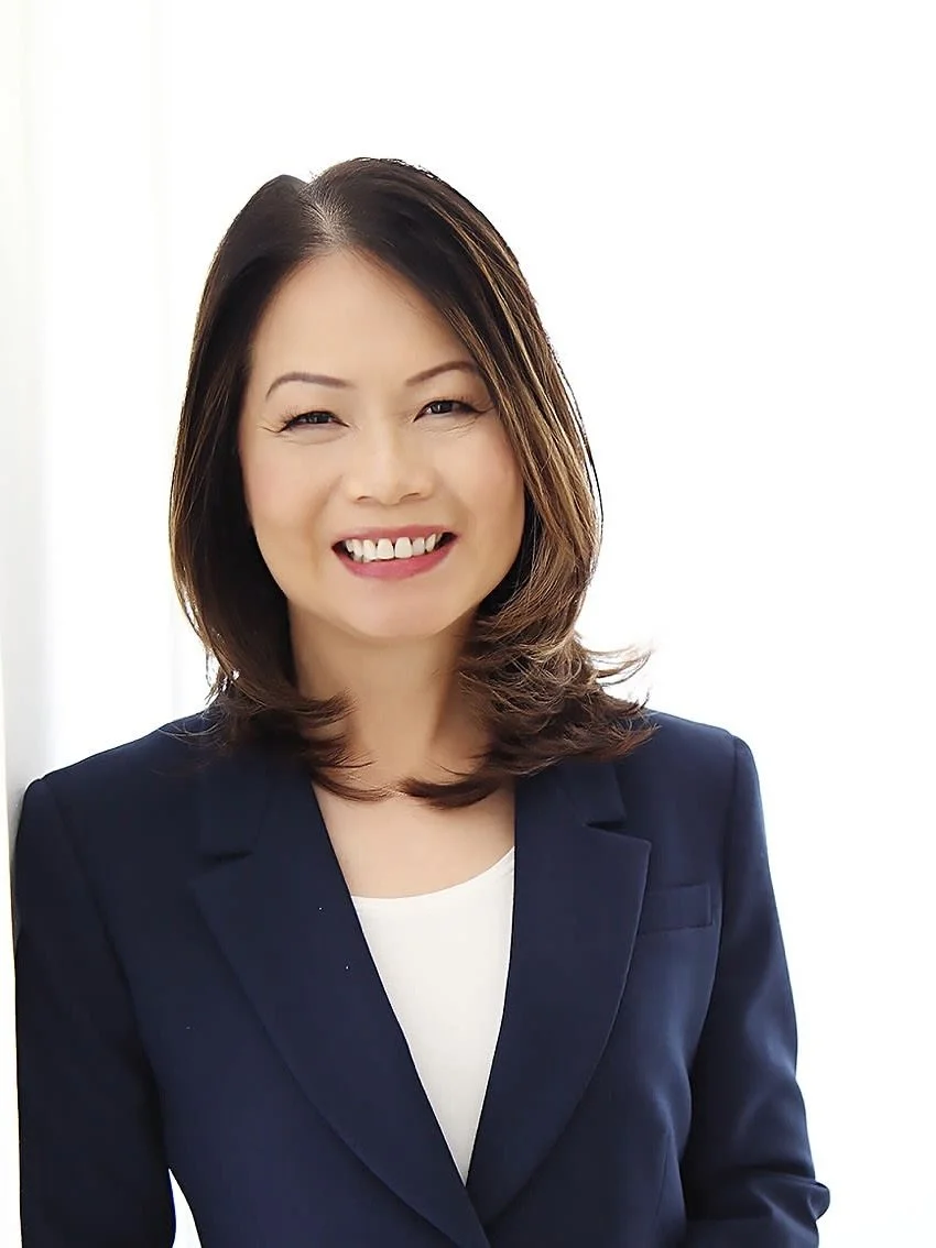 Professional woman with shoulder-length dark hair smiling in a navy blazer and white blouse, standing against a white background.
