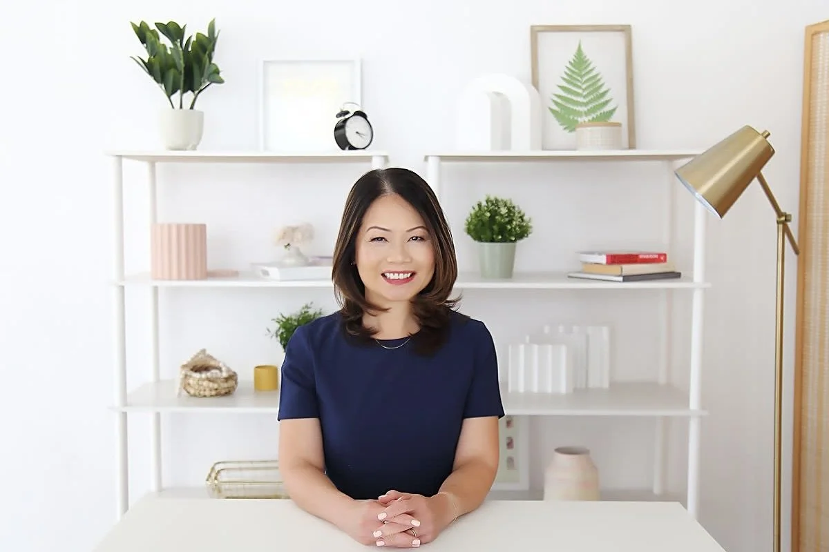A smiling woman with shoulder-length dark hair, wearing a navy blue top, sitting at a white table in a bright, decorated room with shelves holding plants, picture frames, books, and decorative items.